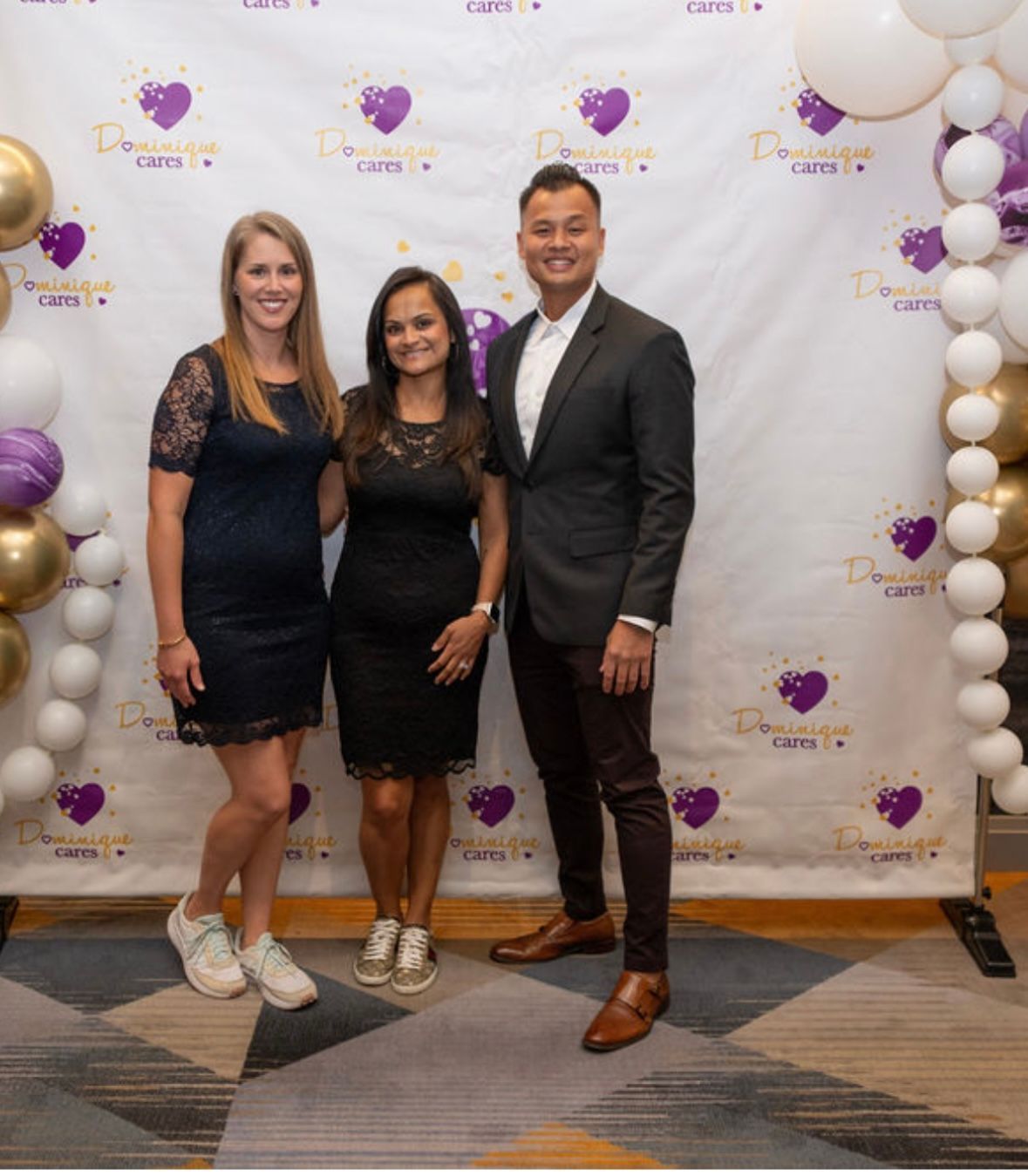 Three people pose in front of a backdrop with balloons. Woman in navy dress, woman in black dress, man in gray suit.