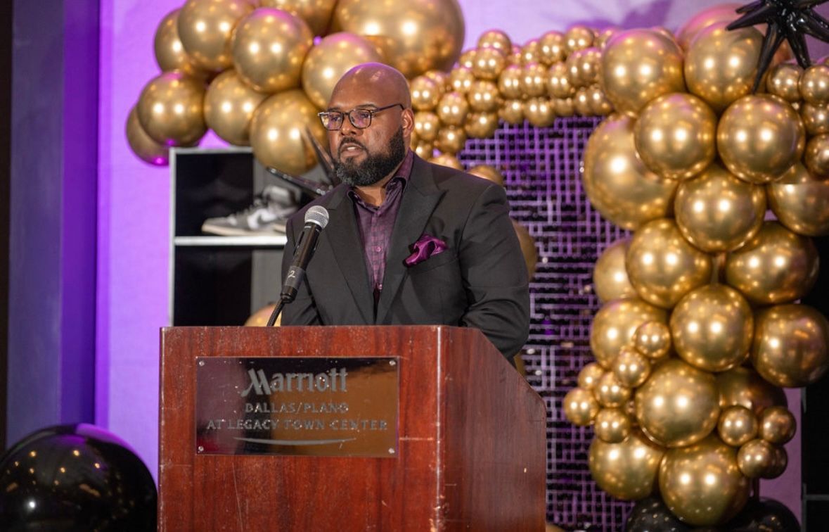 Man speaking at a podium with a Marriott logo, gold balloons, and purple accents.