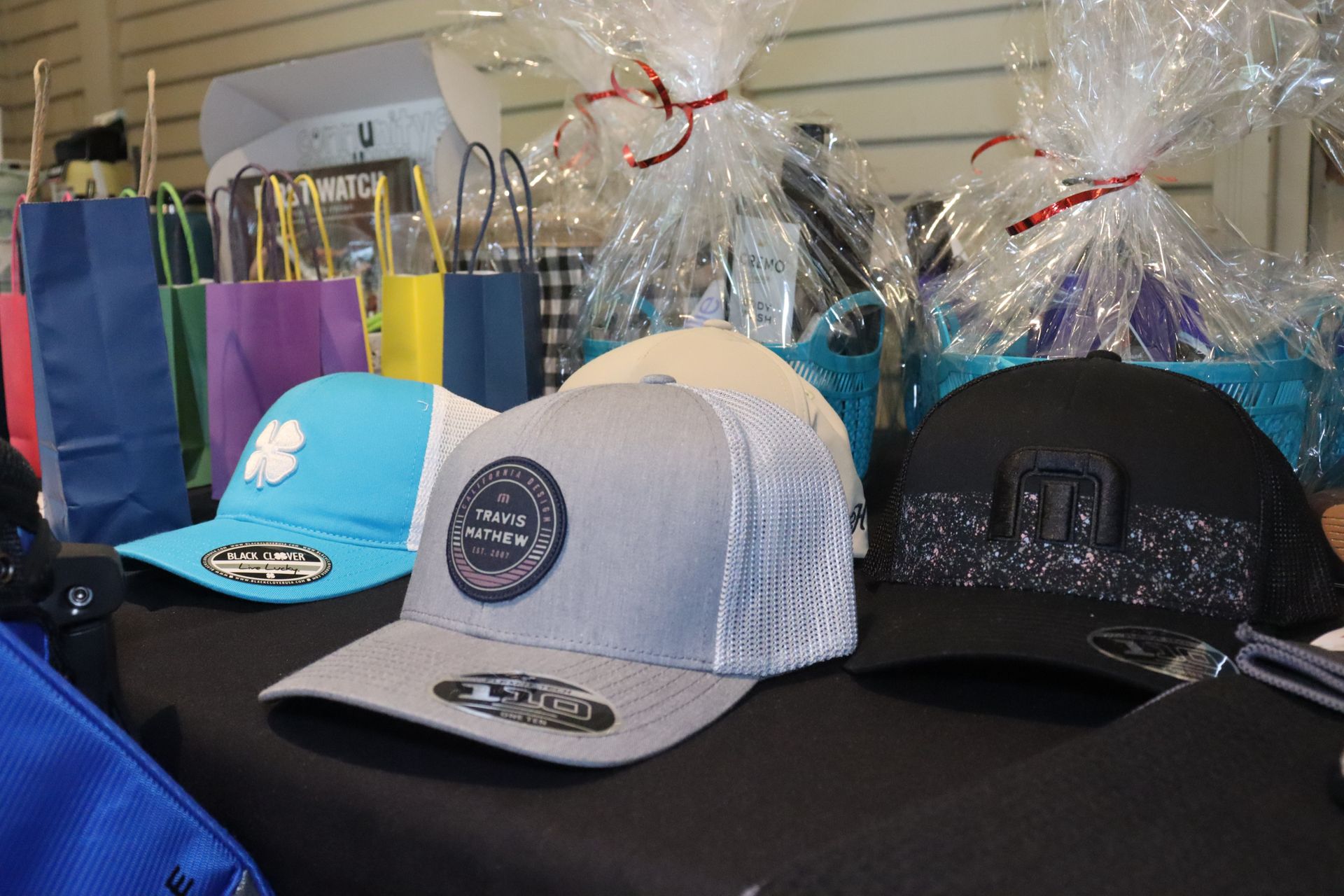 Caps for sale on a table. Gray, blue, and black hats with logos, gift baskets, and shopping bags.