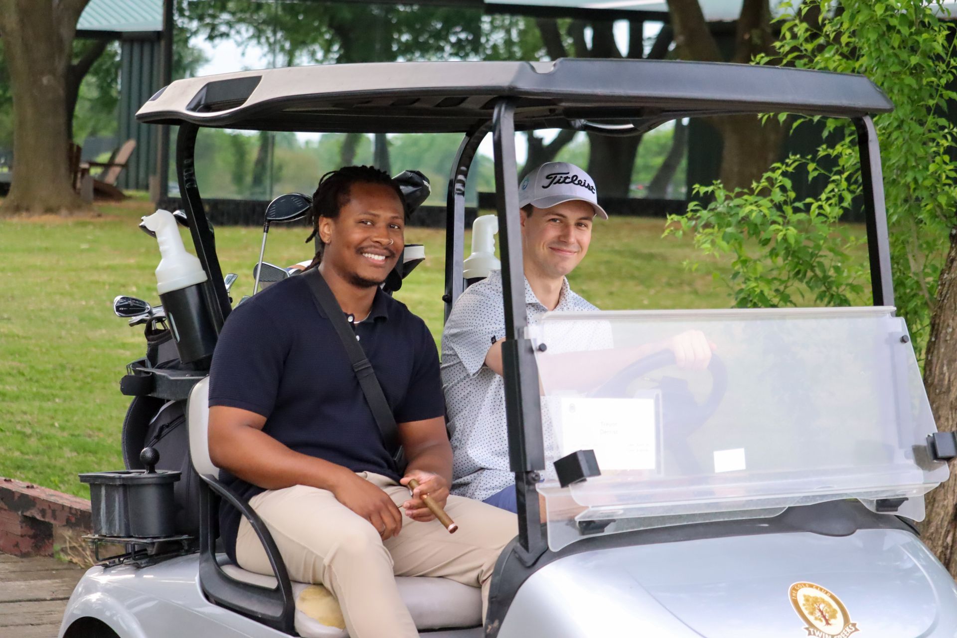 Two men smiling in a silver golf cart on a green course; one holds a cigar.