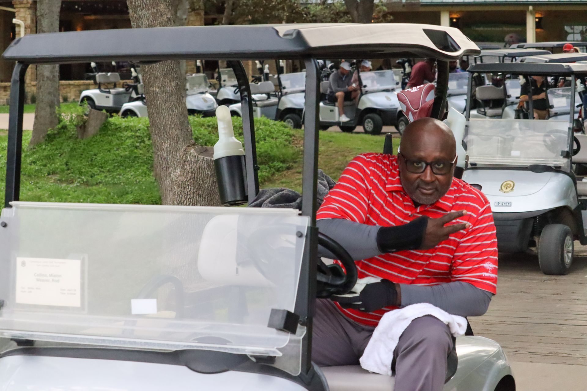 Man in red shirt gestures from golf cart, surrounded by others at a golf course.