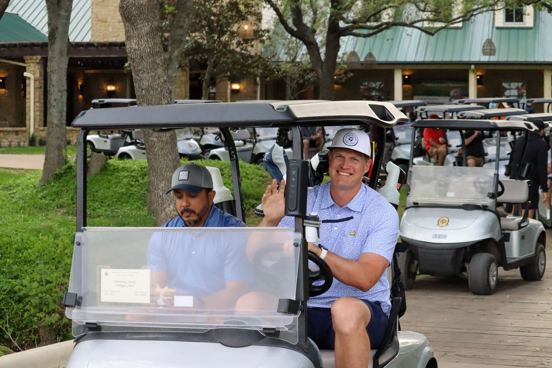 Men in golf carts, waving and smiling, near a building and trees.