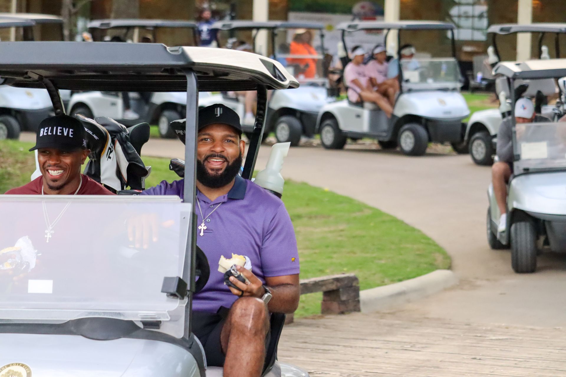 Men in golf carts on a course, smiling. One wears purple shirt. Other carts visible behind them.
