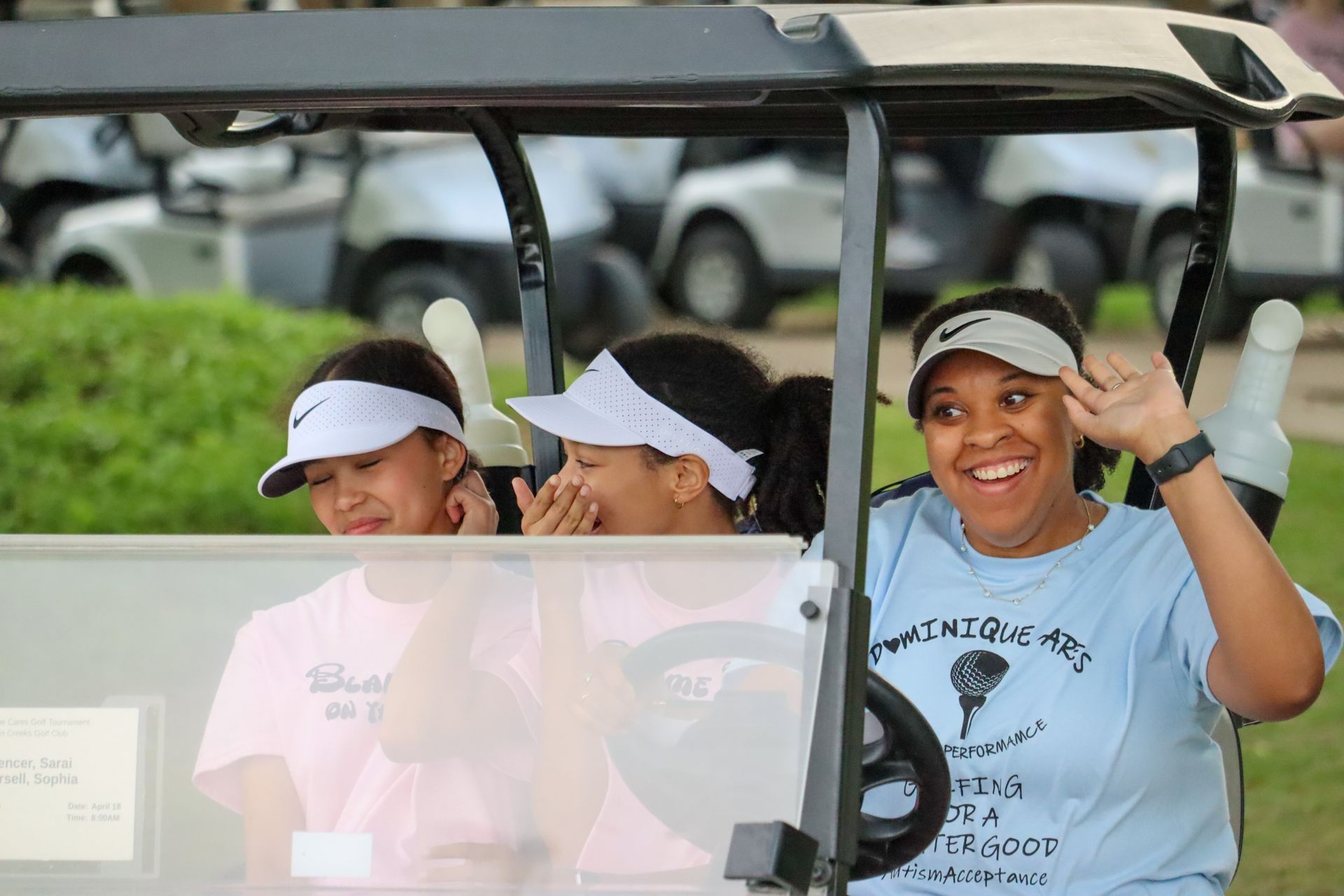 Three women in a golf cart. Two wearing pink, one waving. Sunny day with golf carts in background.