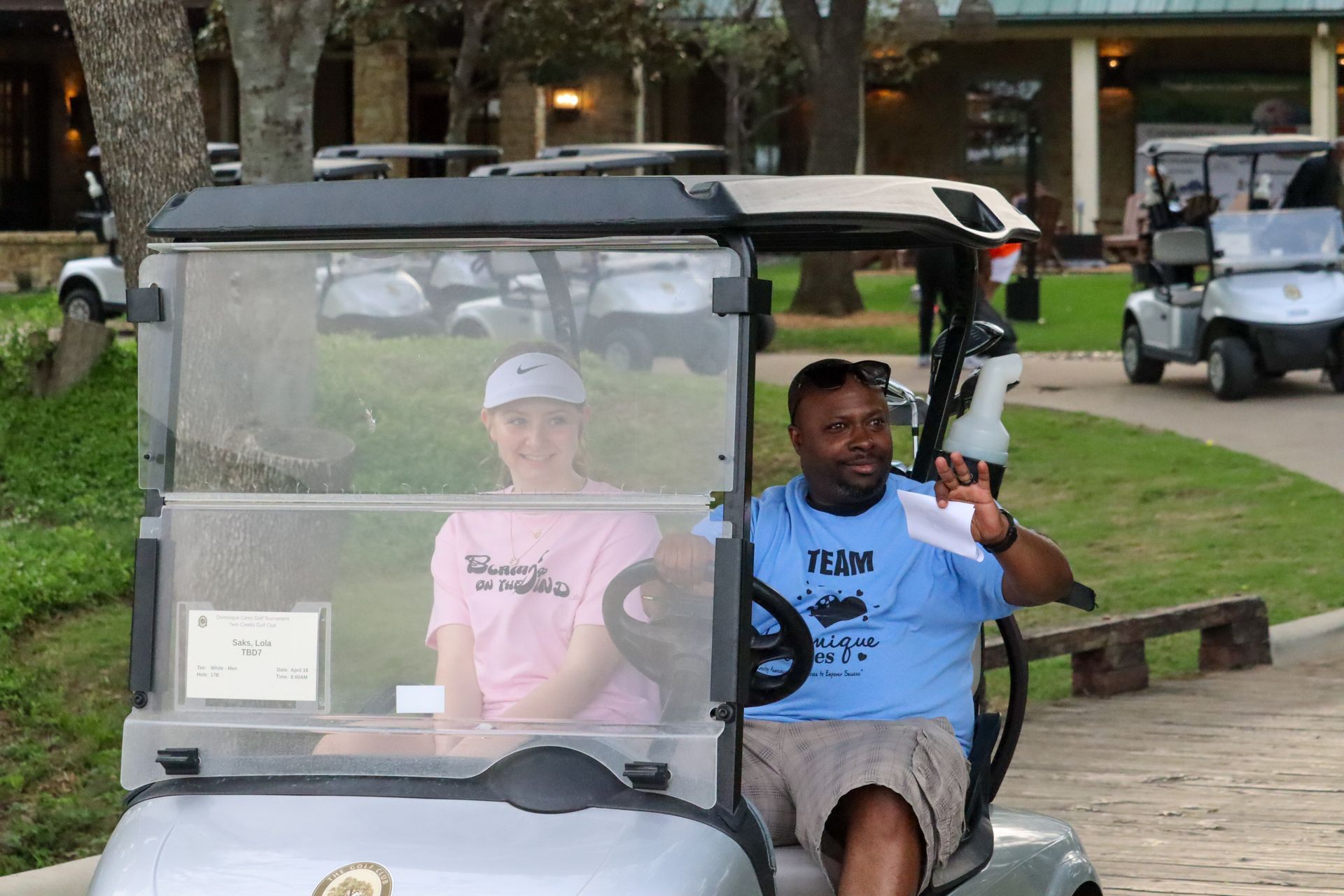 Two people in a golf cart; one waves, other smiles. Daytime, outdoor setting with other carts and buildings.
