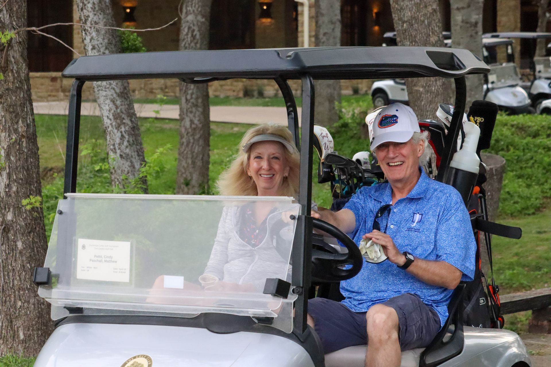 A smiling couple in a golf cart on a course. Man holds money, woman smiles. Trees and other carts visible.