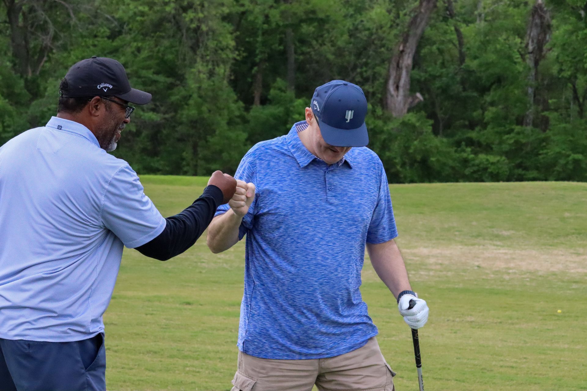 Two men on a golf course: one fist-bumps the other, who holds a club. Green grass, blue shirts.