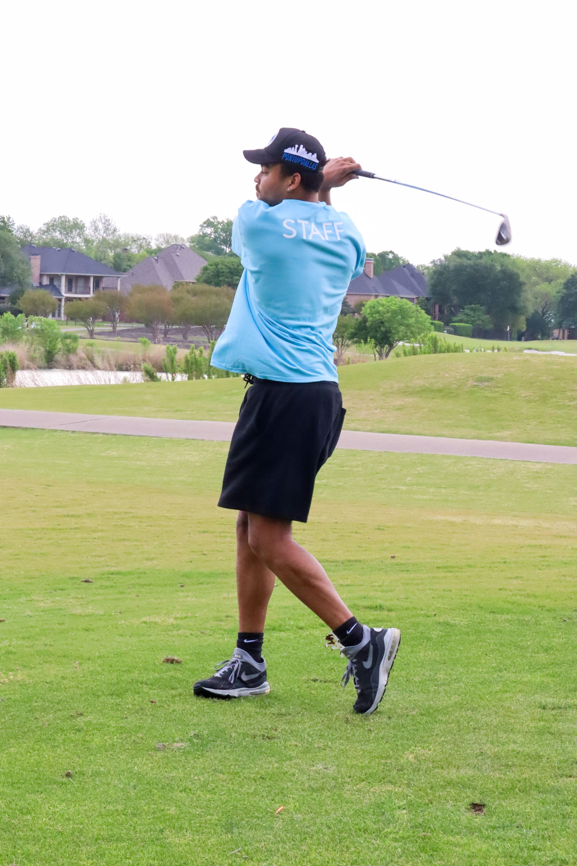 Golfer in blue shirt and black shorts swinging club on green golf course.