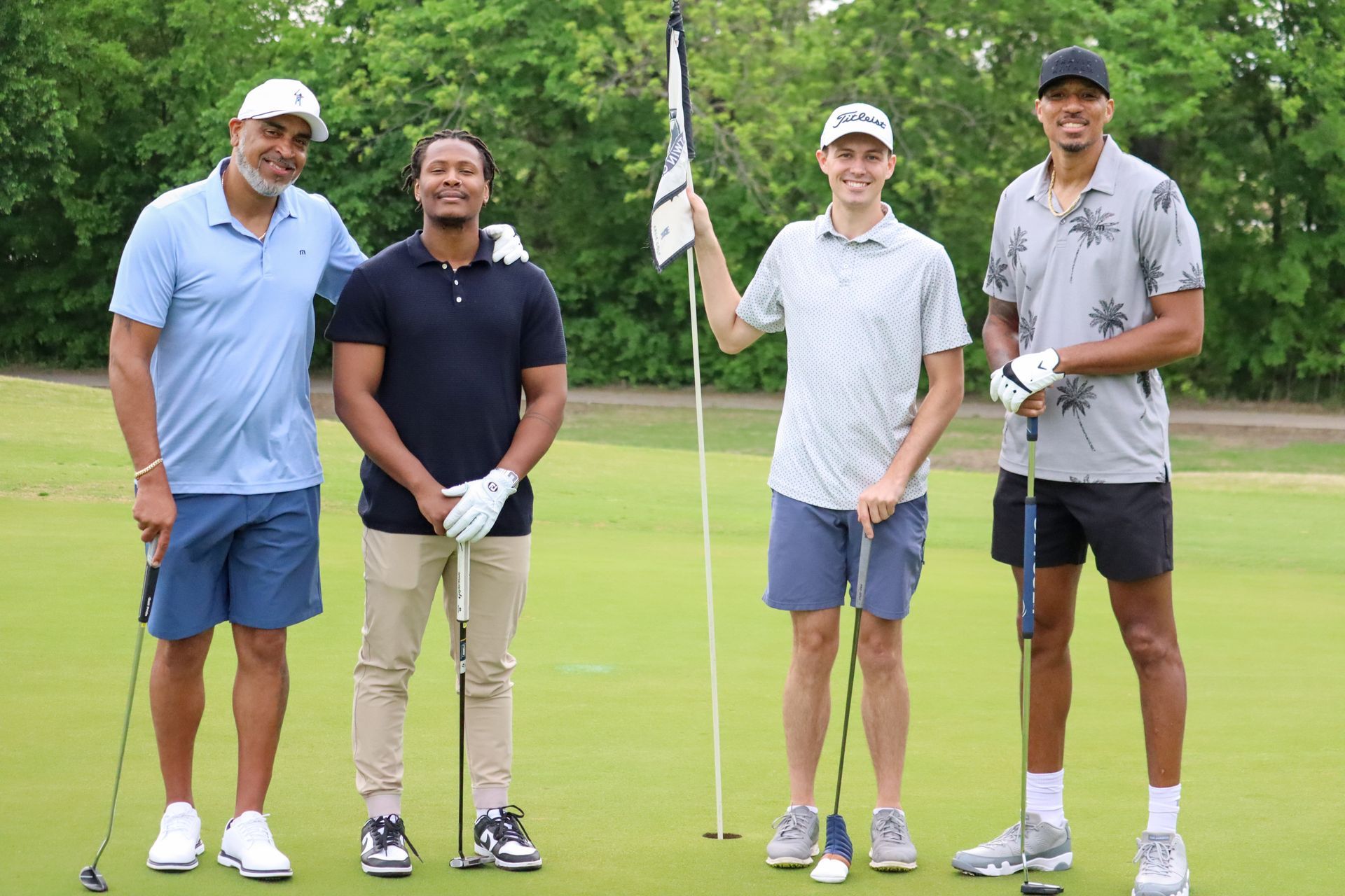 Four men on a golf course pose with clubs, smiling near a flag.