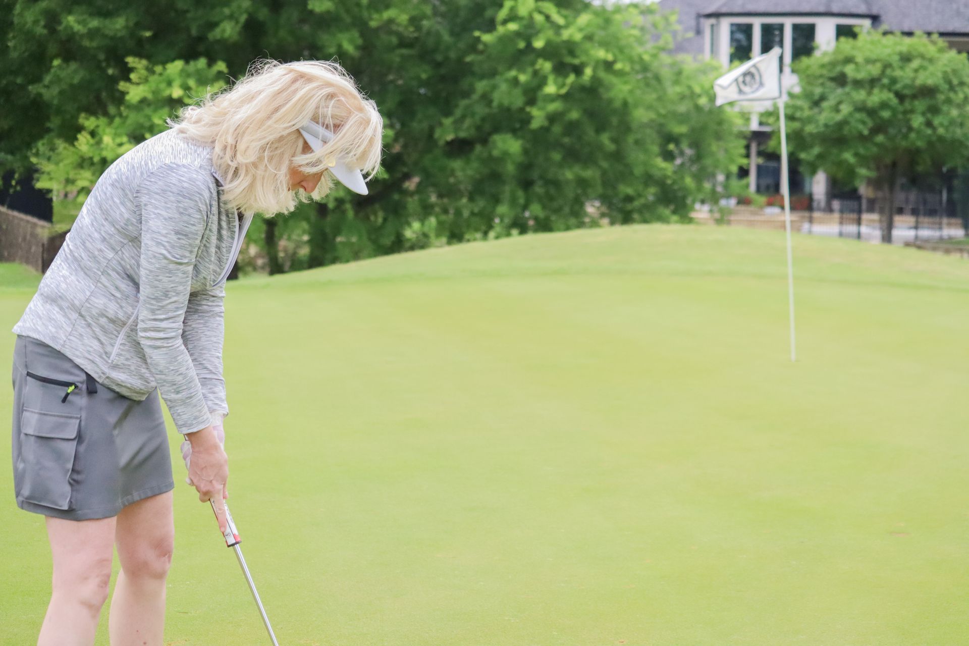Woman putting golf ball on a green; wearing visor, grey top, and shorts. Golf course in the background.