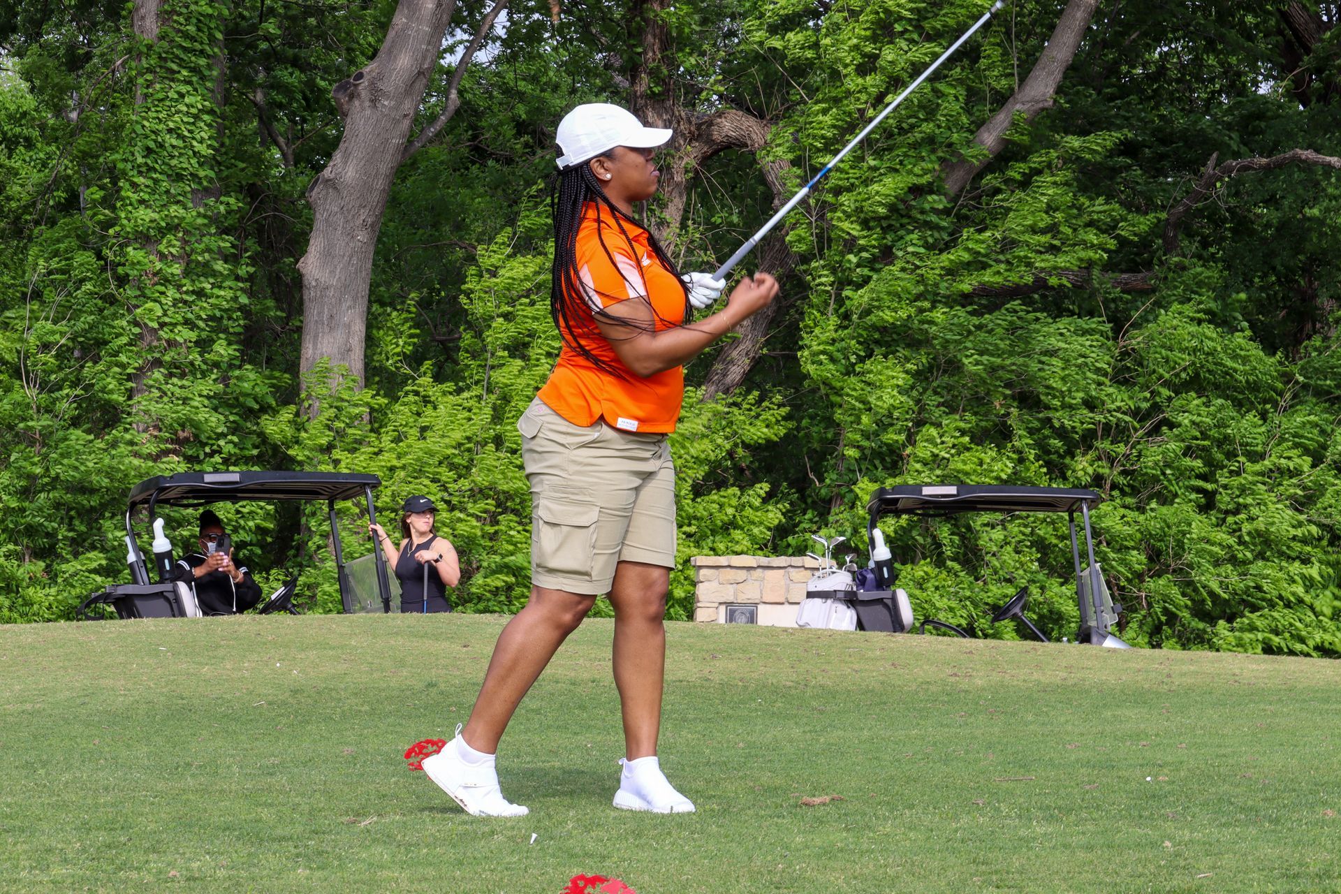 Woman in orange shirt swings a golf club on a green course. Trees and golf carts in background.