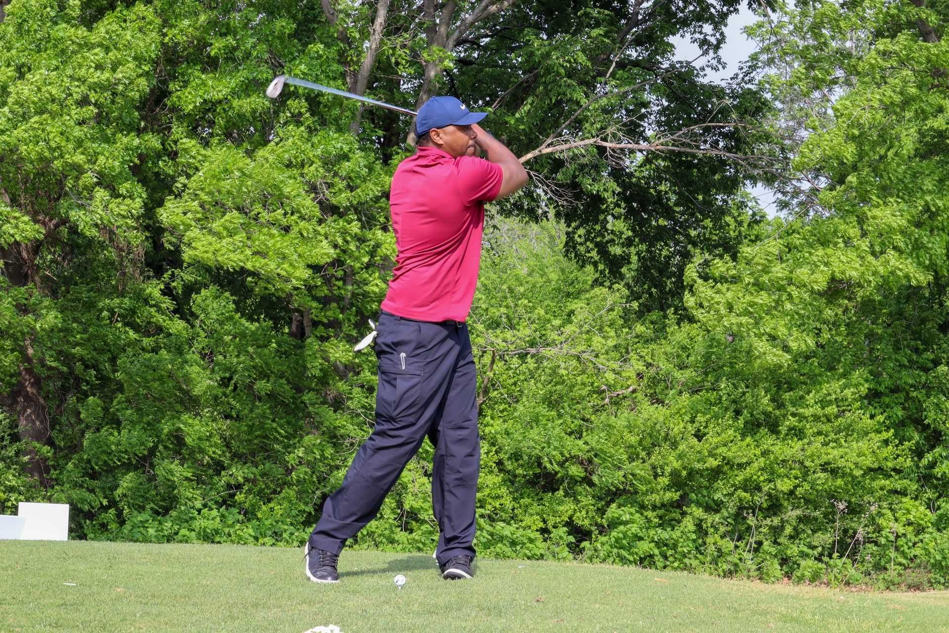 Golfer in red shirt and blue hat swings club on a green tee box, surrounded by trees.