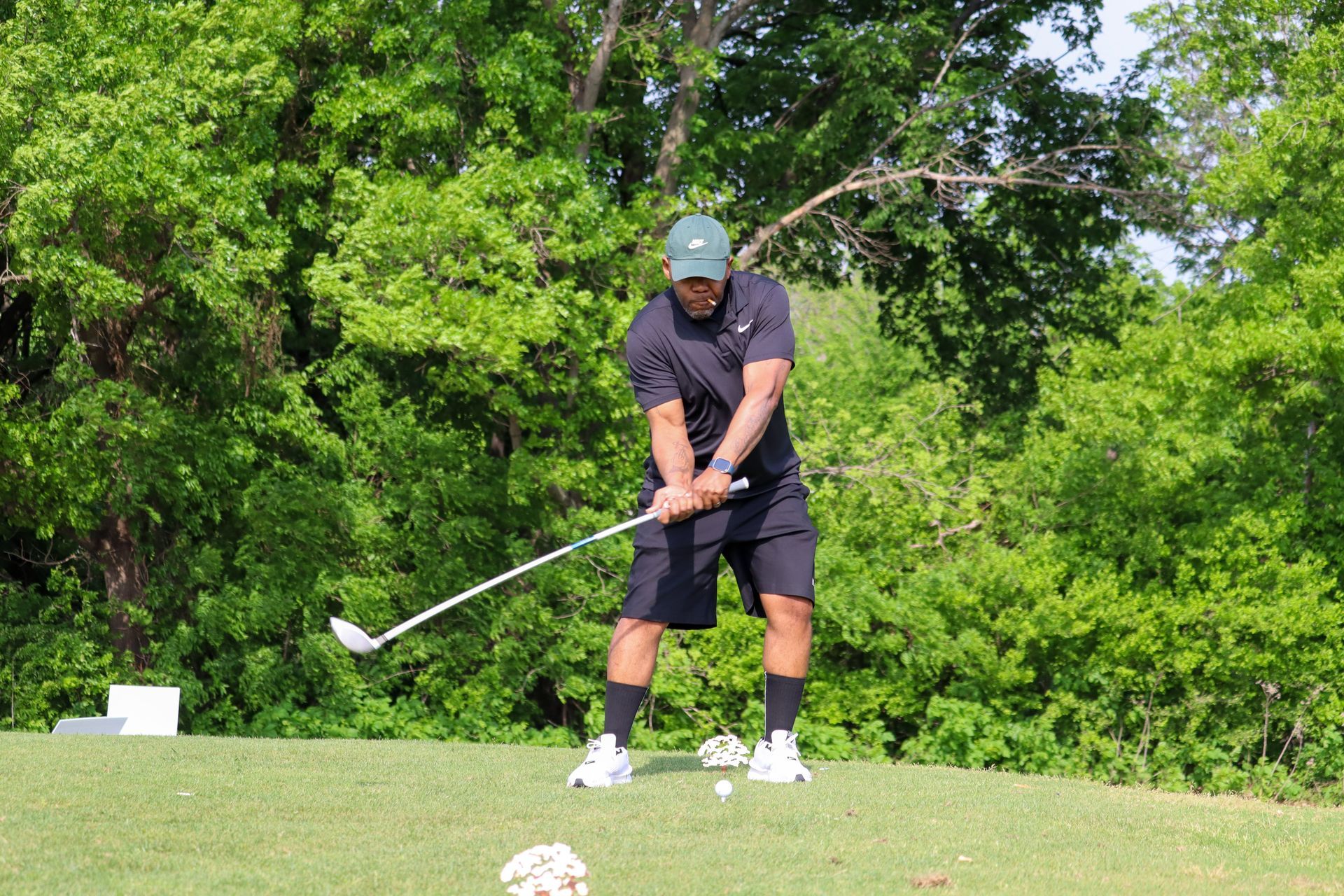 Man in black golf attire swinging a club on a green, surrounded by trees.