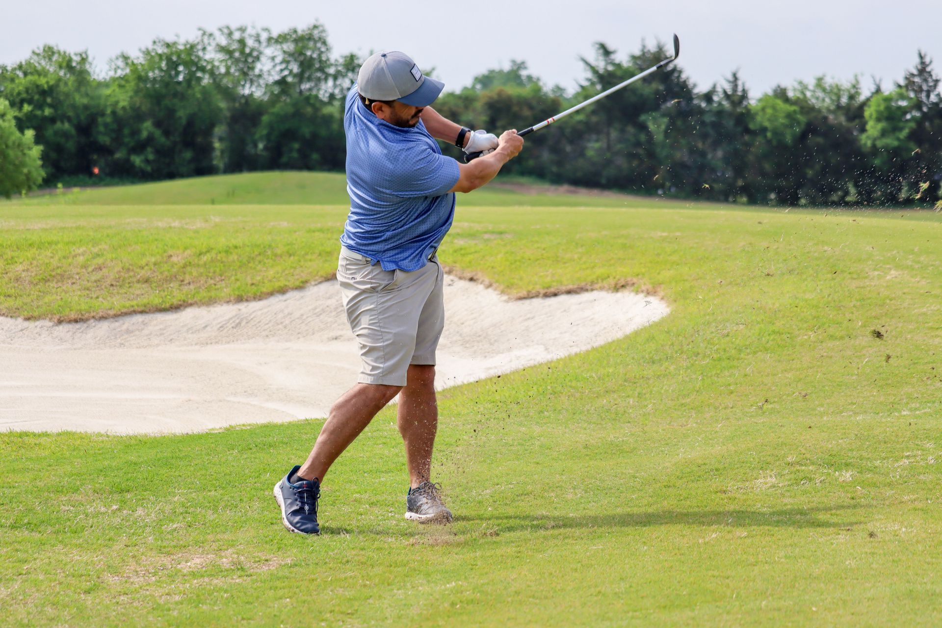 Golfer swinging a club on a green, near a sand trap, wearing a hat and blue patterned shirt.