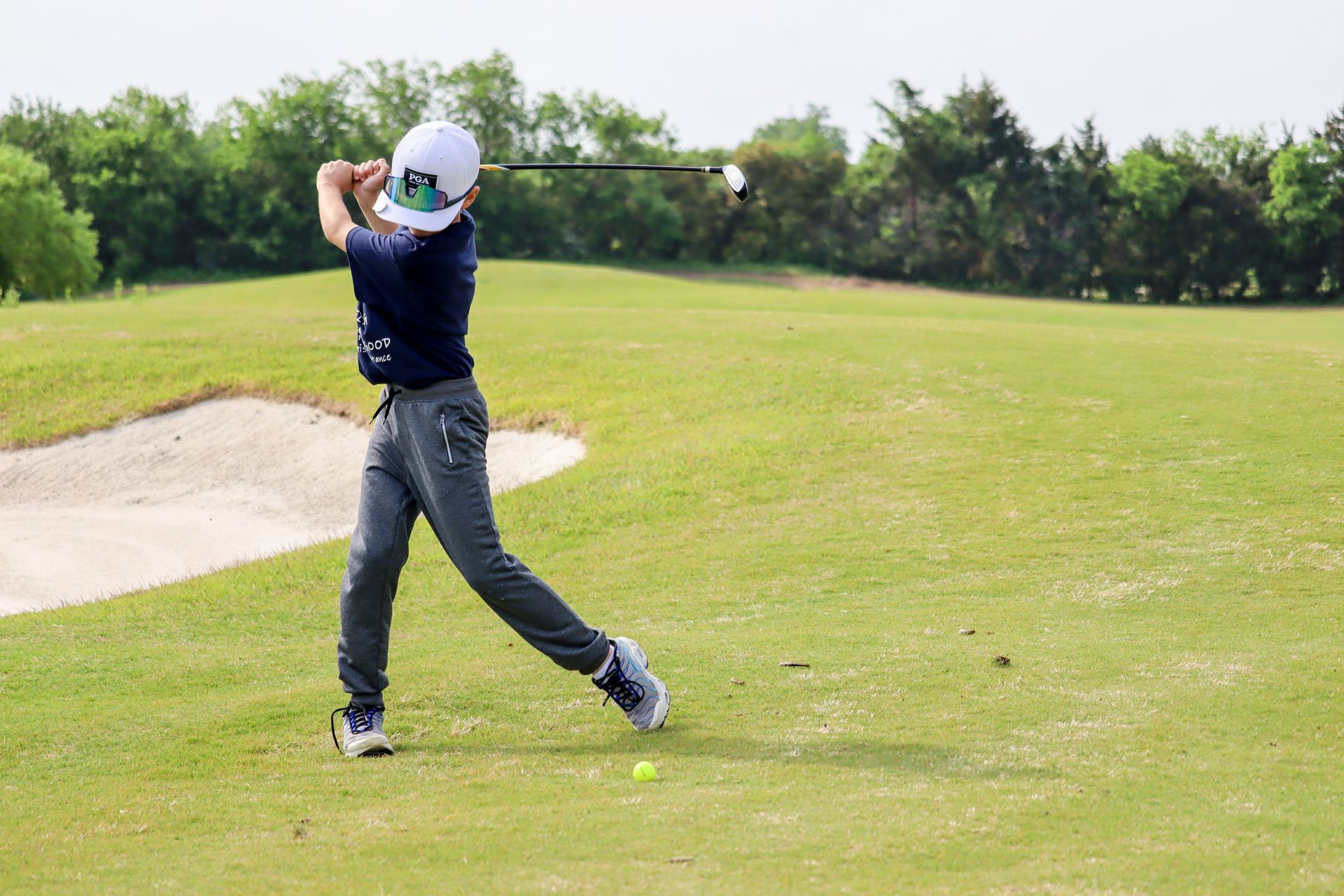 Boy in a baseball cap swinging a golf club on a green course; a sand trap is visible.