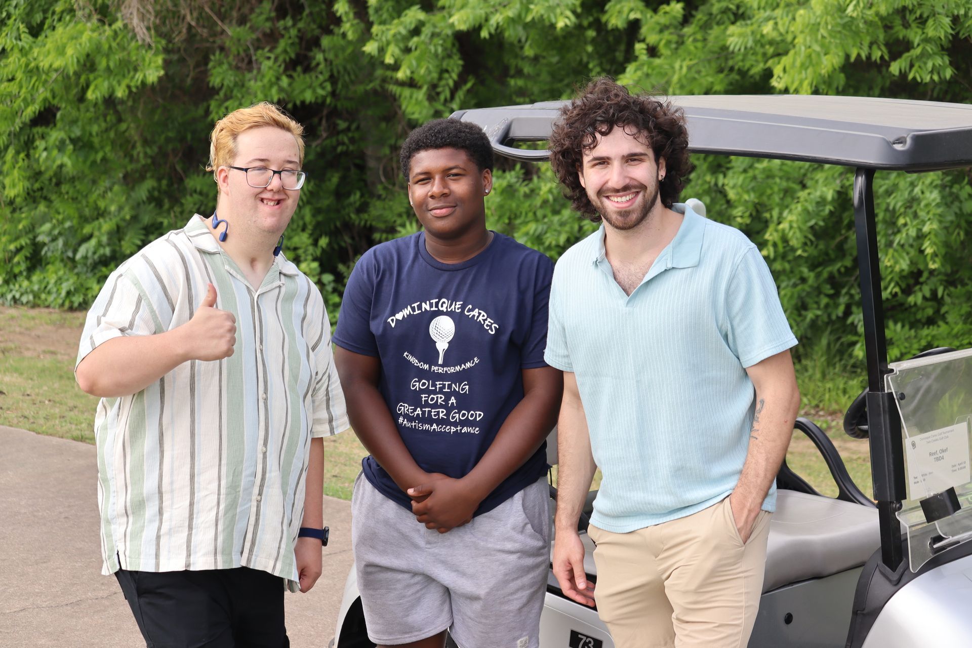 Three men pose by a golf cart. One gives a thumbs up. Outdoors, trees in background.