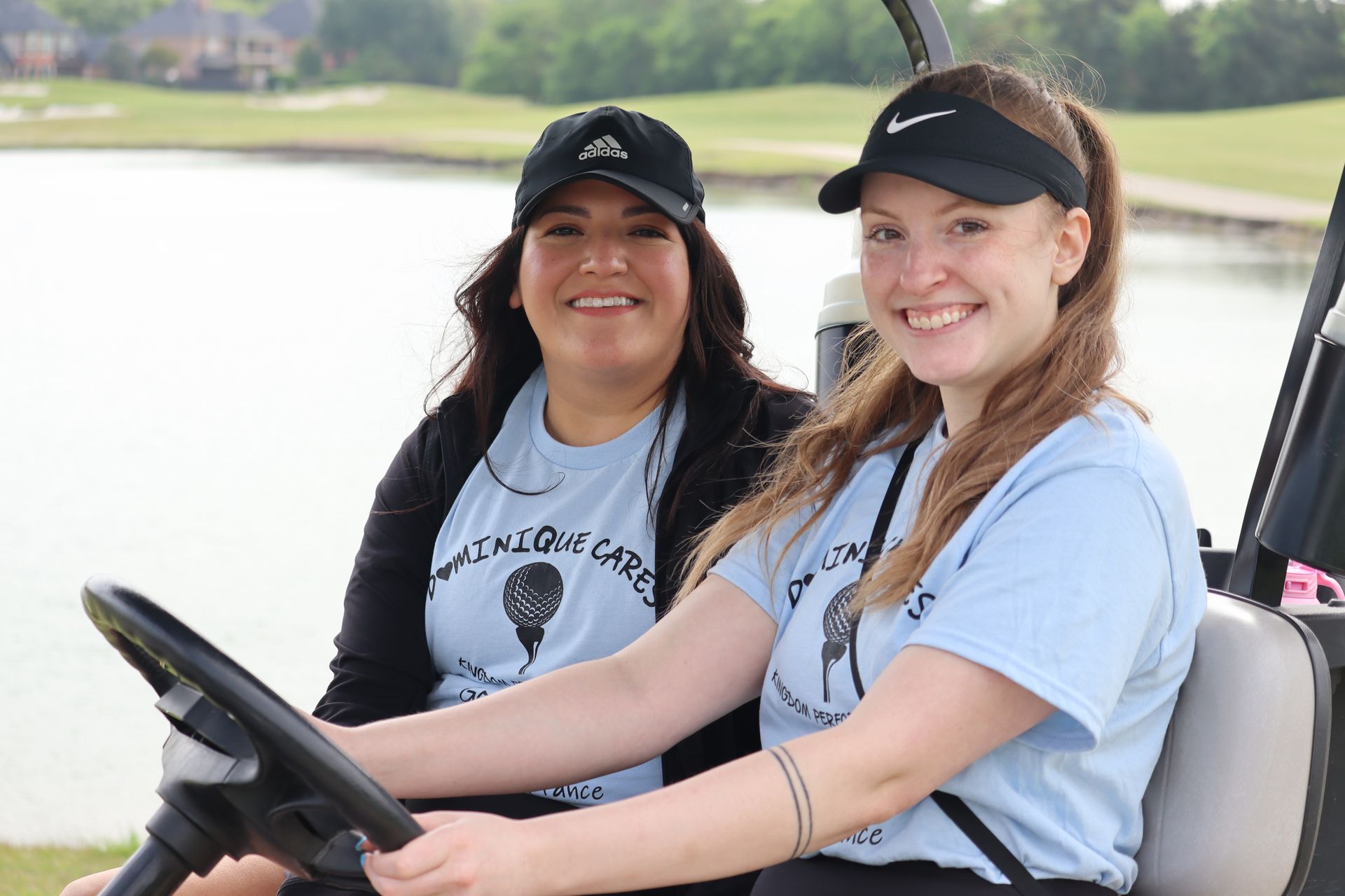 Two women in a golf cart smile at the camera, wearing light blue shirts and hats with a golf course behind them.