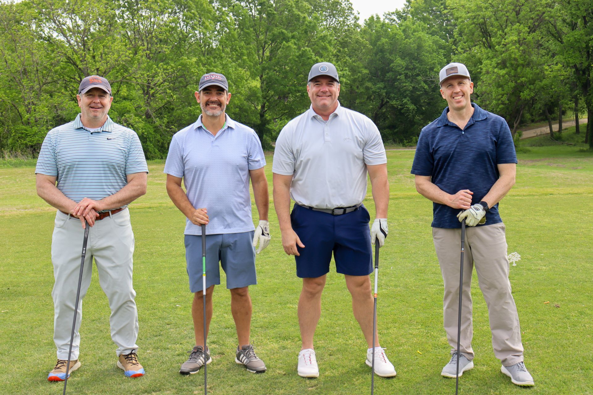 Four men on a golf course, each holding a golf club, smiling and posing in casual attire.