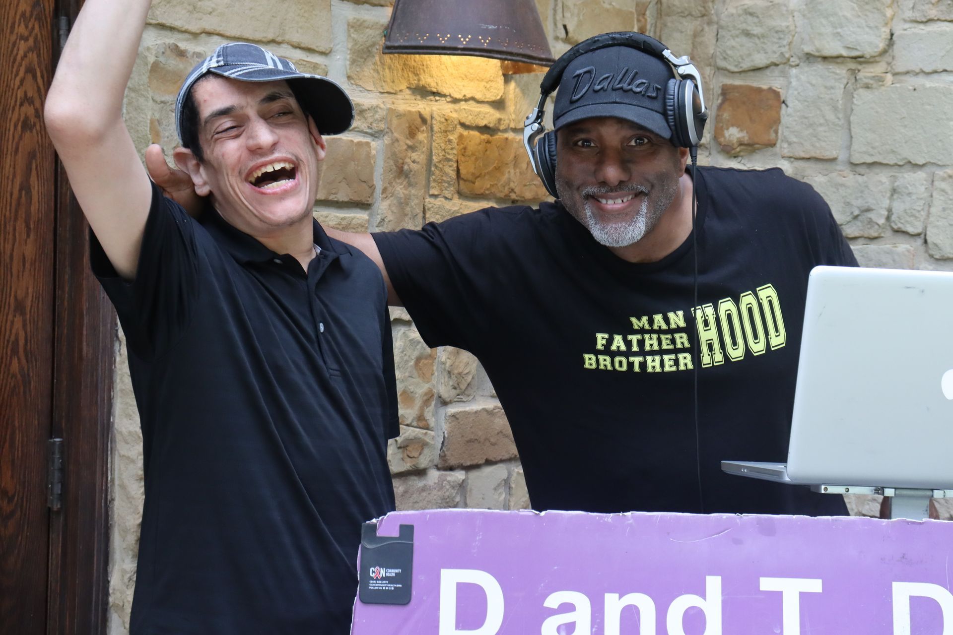 Two men smiling by DJ equipment, one in black hat, one with headphones on, at an outdoor event.