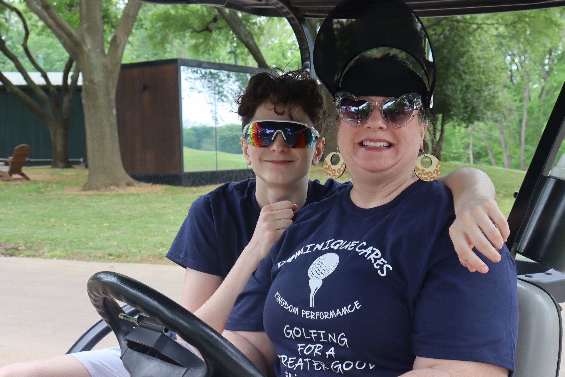 Woman and teen in golf cart, smiling, wearing sunglasses. Outdoors, trees, building in background.