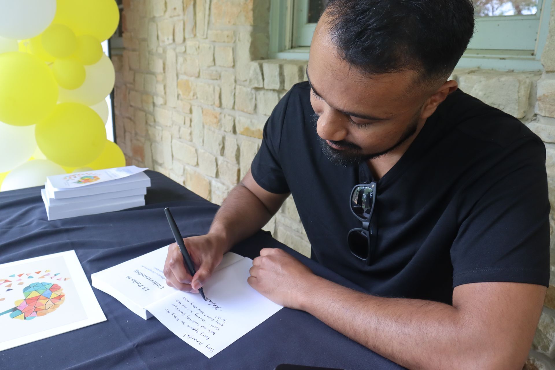 Man in black shirt writing at a table; balloons in background.
