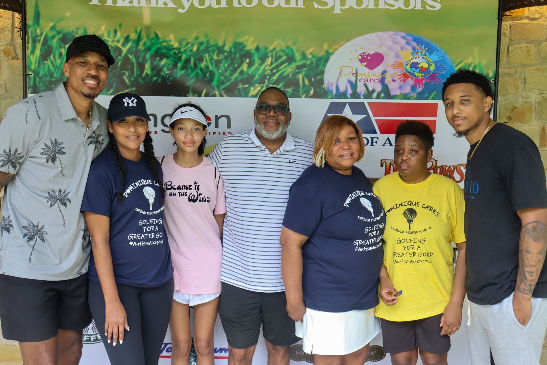 Group of people smiling at a golf outing. Green grass backdrop with sponsor logos.