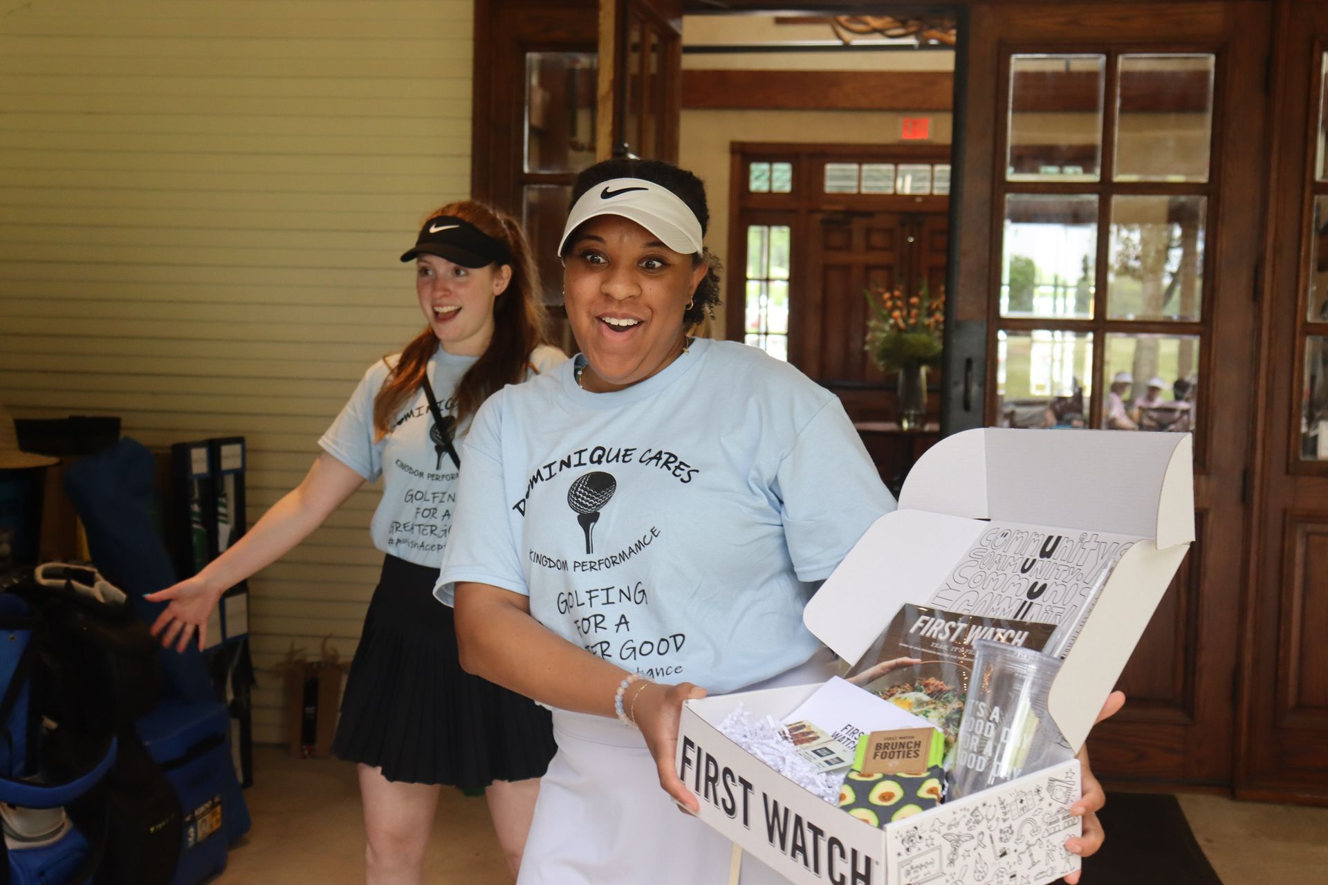 Two women in matching light blue shirts holding a box; golf course exterior.