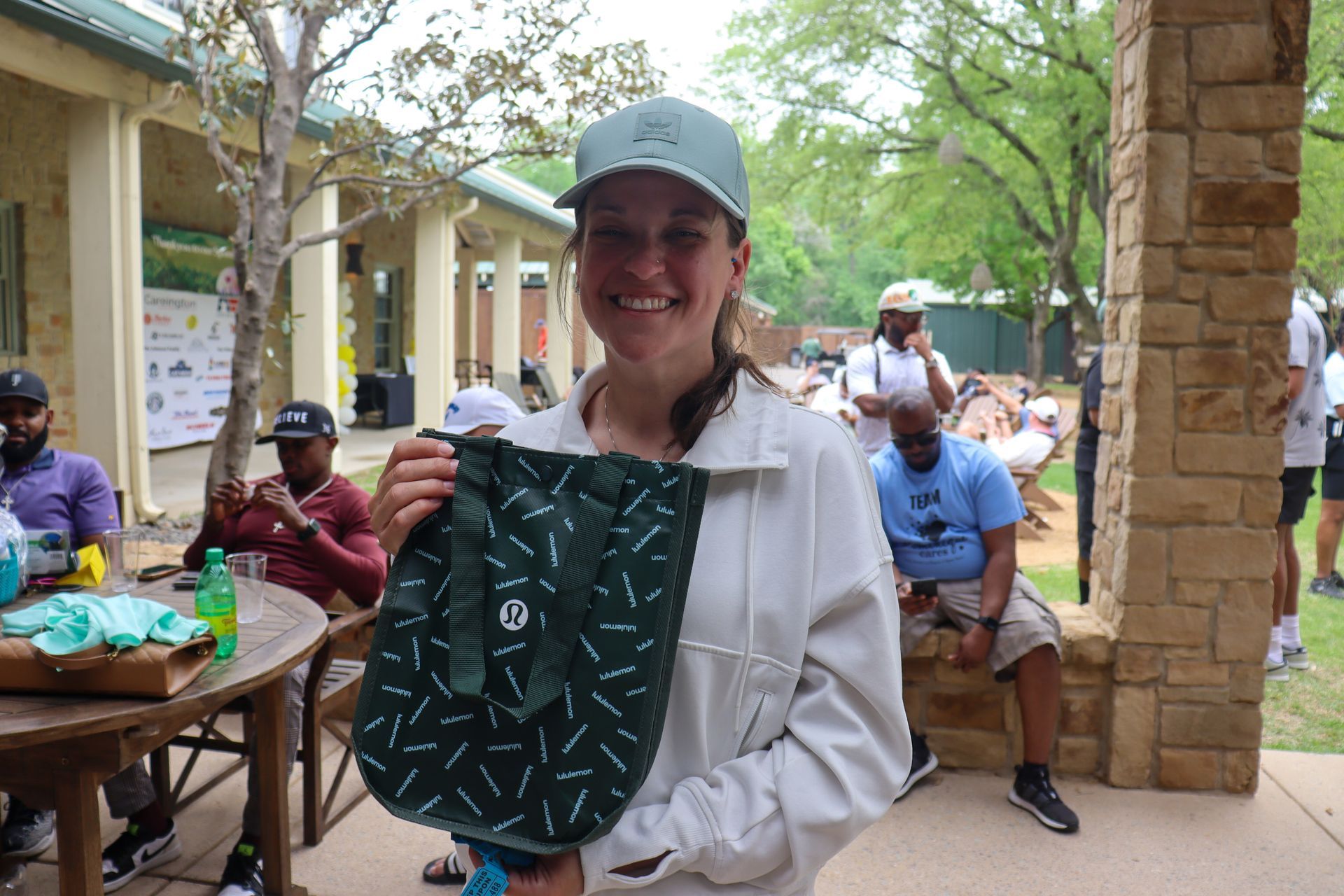 Woman in cap holding a green bag, smiling outdoors near a table with people.