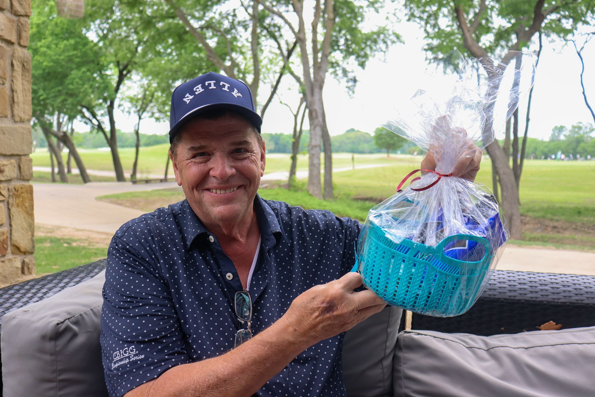 Man smiles, holding a blue Easter basket on an outdoor patio overlooking a golf course. He wears a navy hat and shirt.