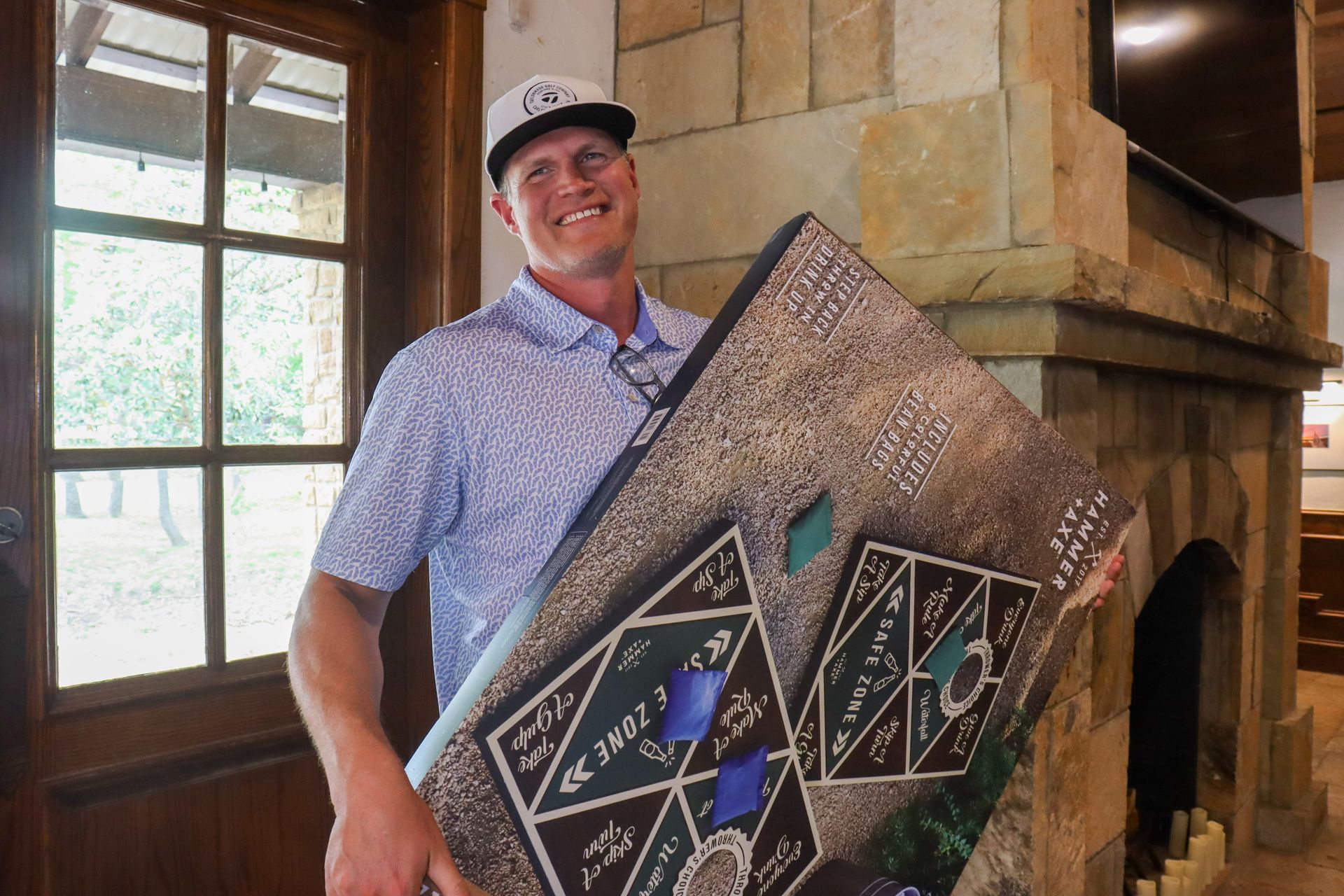 Man smiles, holding decorated game board by fireplace. White cap, blue shirt.