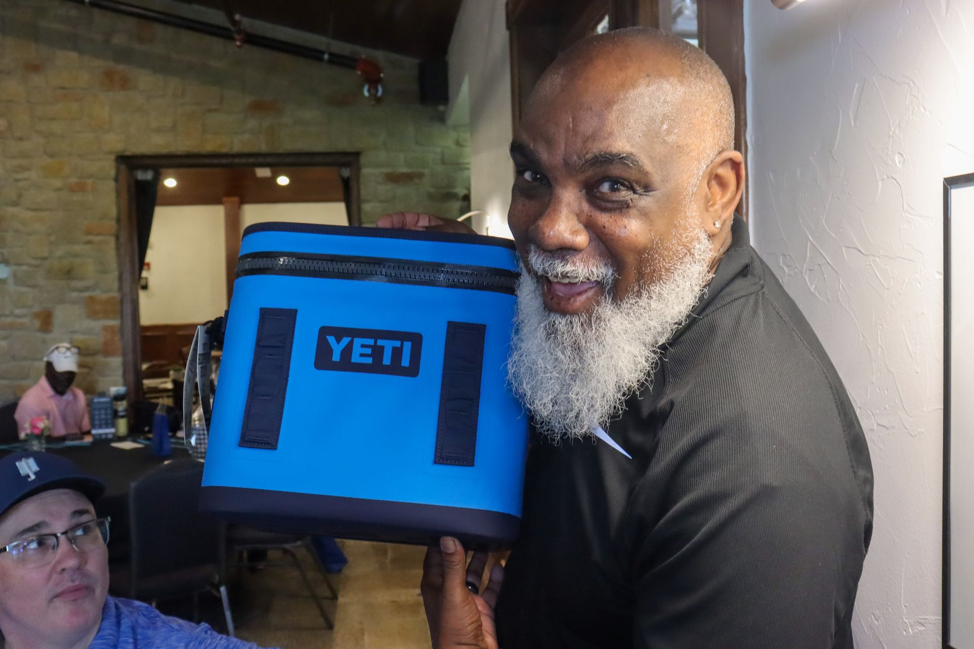 Man holding a blue Yeti cooler; smiling indoors, another person looking at the camera.
