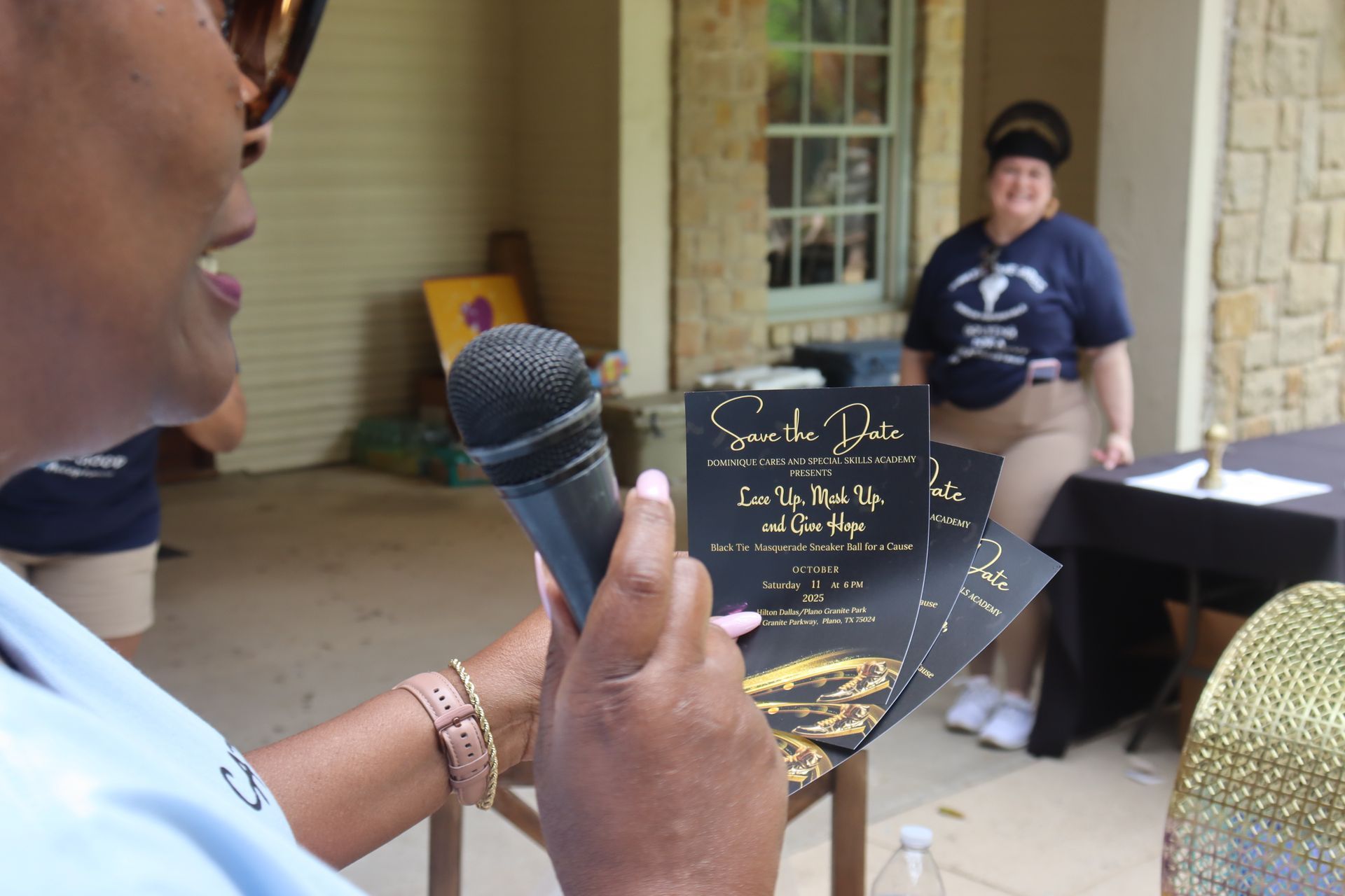 Woman speaking into a microphone, holding event flyers. Another woman in background. Outdoors, day.