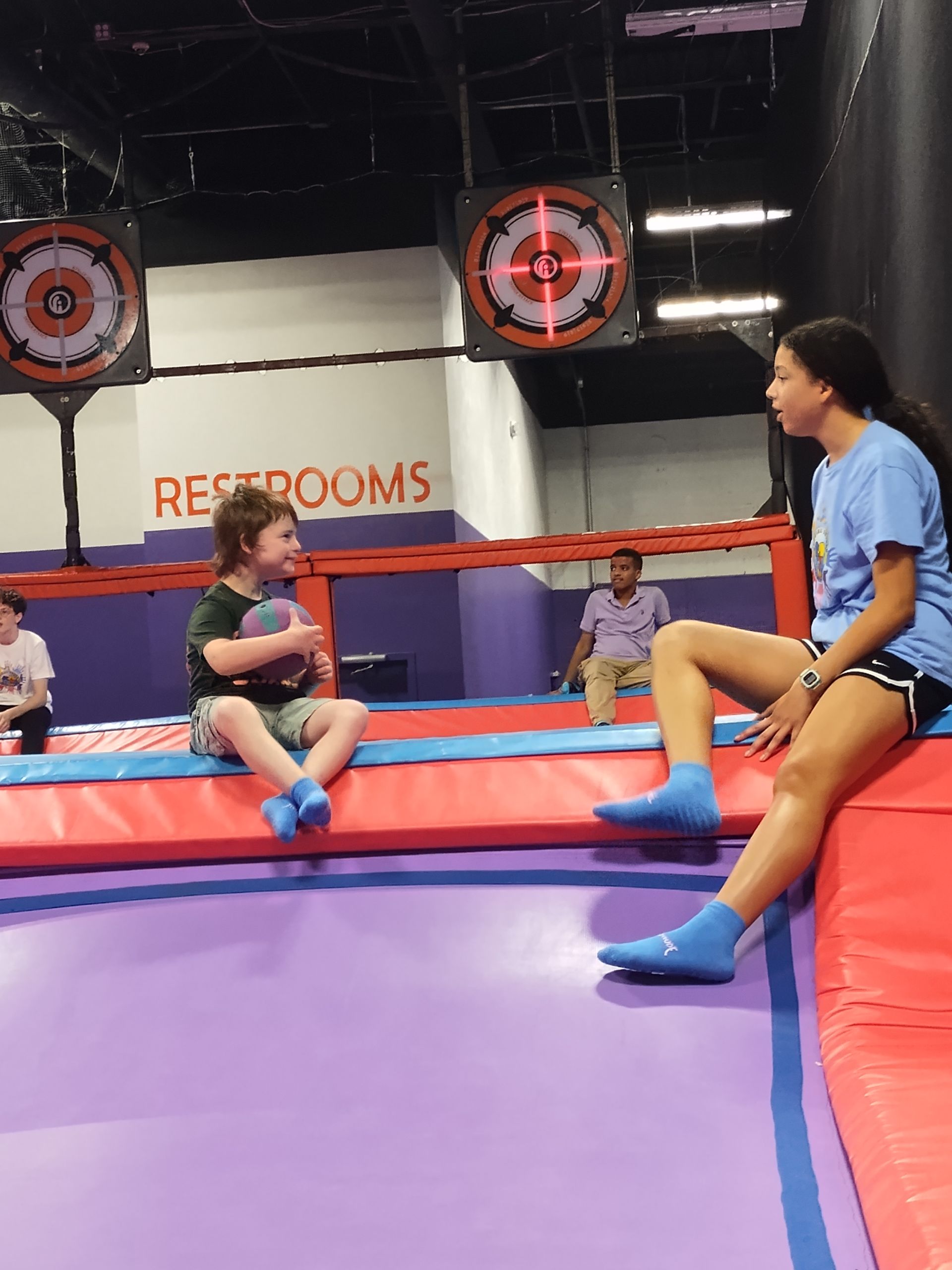 Two people, one holding a ball, on a trampoline; a target game in a recreation center.