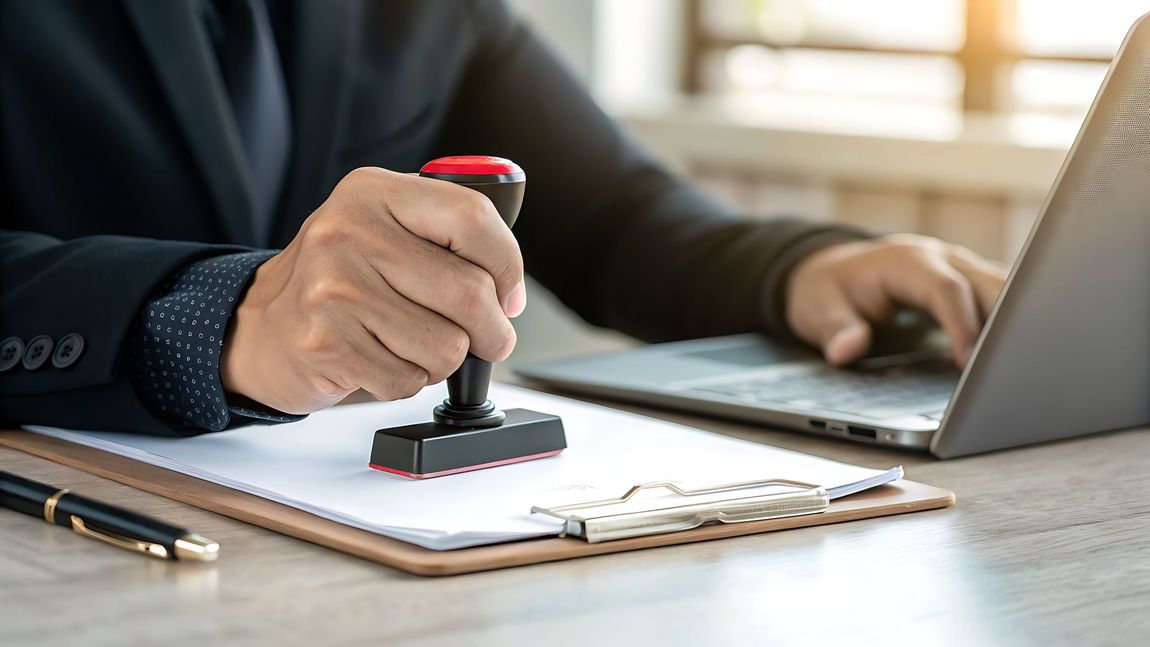 Person in suit stamps document with a red stamp while using a laptop at a desk.