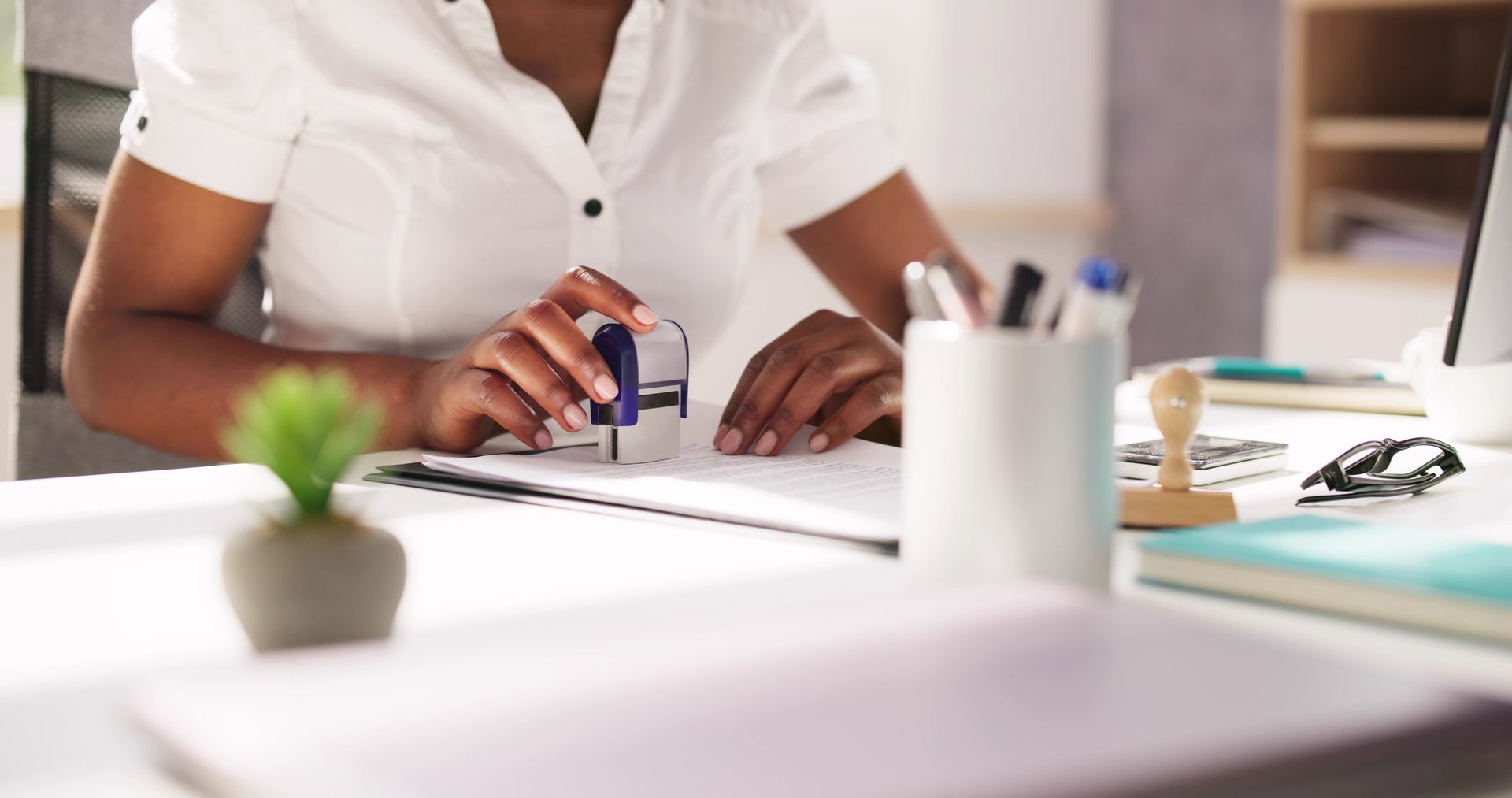 Person stamping a document at a desk; small plant and office supplies nearby.