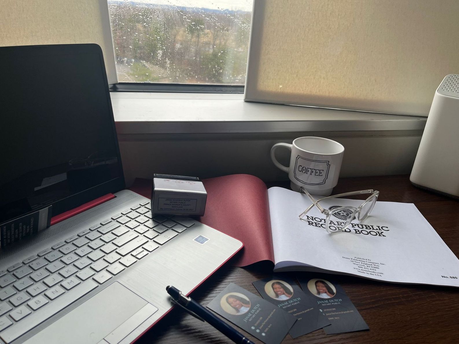 Laptop, mug, open book, business cards, and pen on a desk near a window on a rainy day.