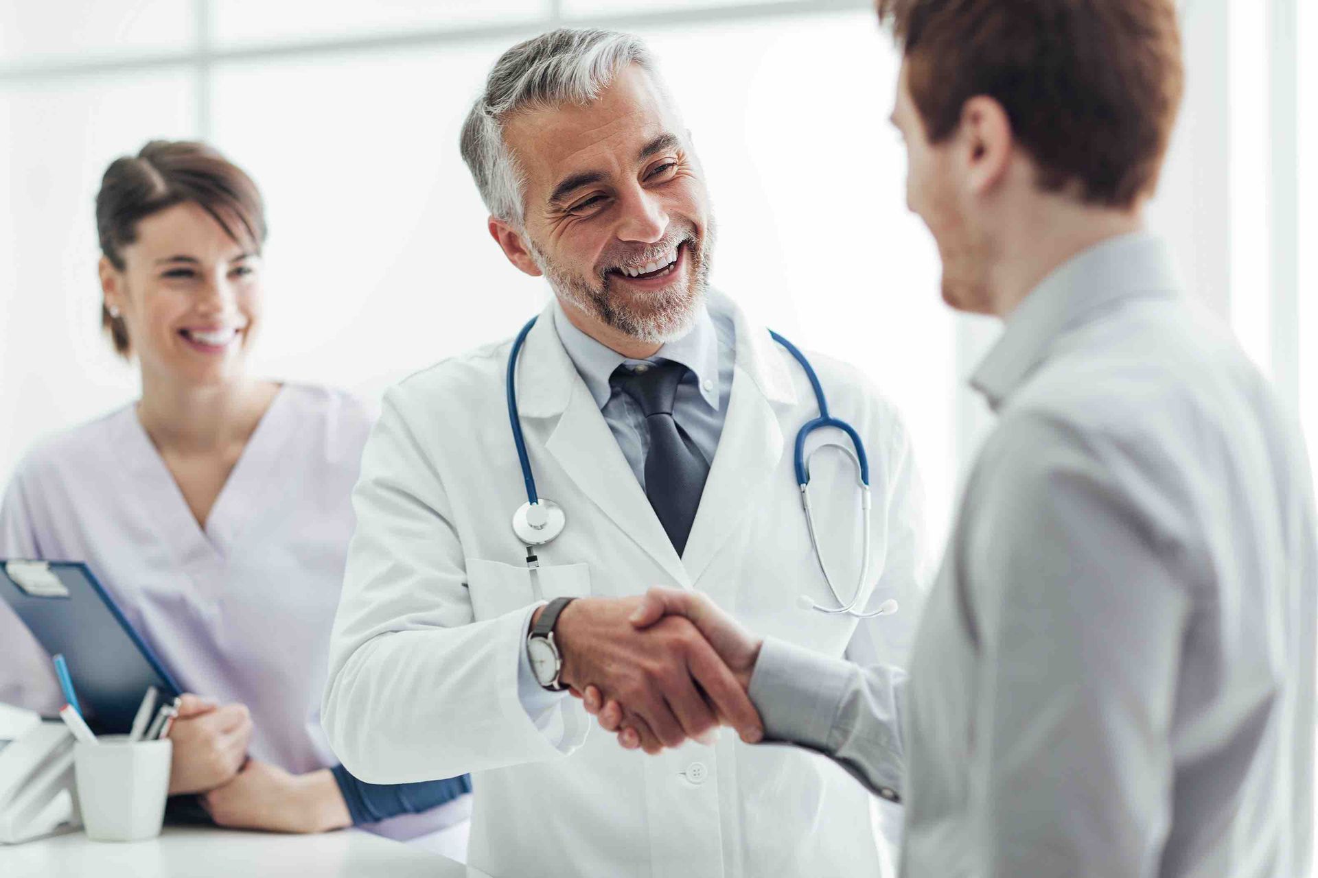 Doctor shaking patient's hand, smiling. Nurse with clipboard in the background, smiling. Clinic setting.