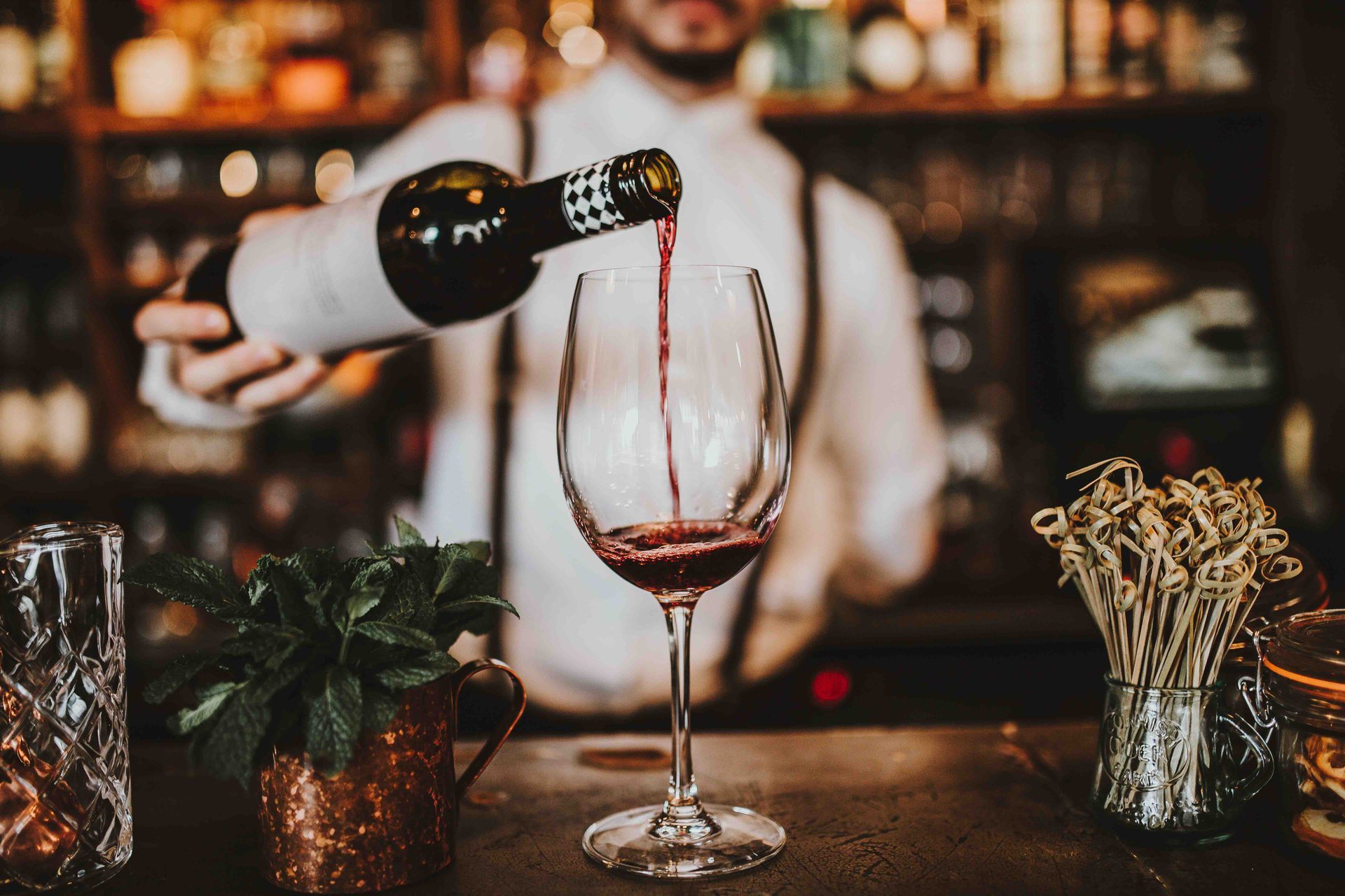 Bartender pouring red wine into a tall glass at a dimly lit bar.