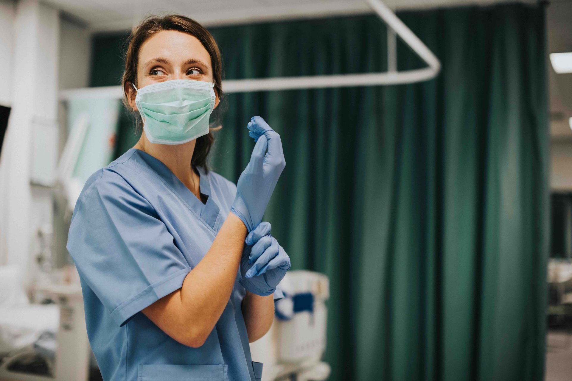 Nurse in blue scrubs and mask putting on gloves in a hospital room.