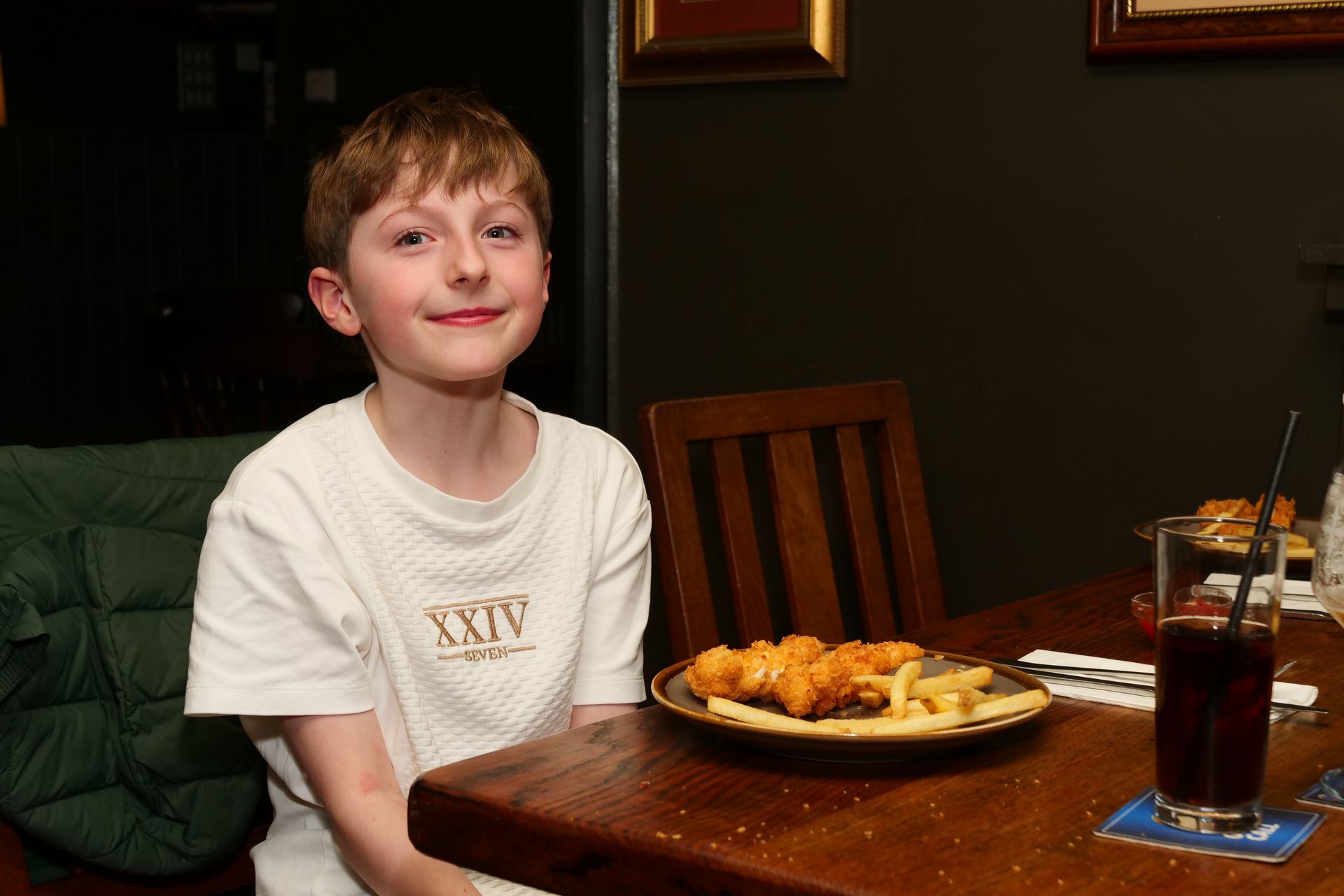 A young boy wearing a xiv shirt sits at a table with a plate of food