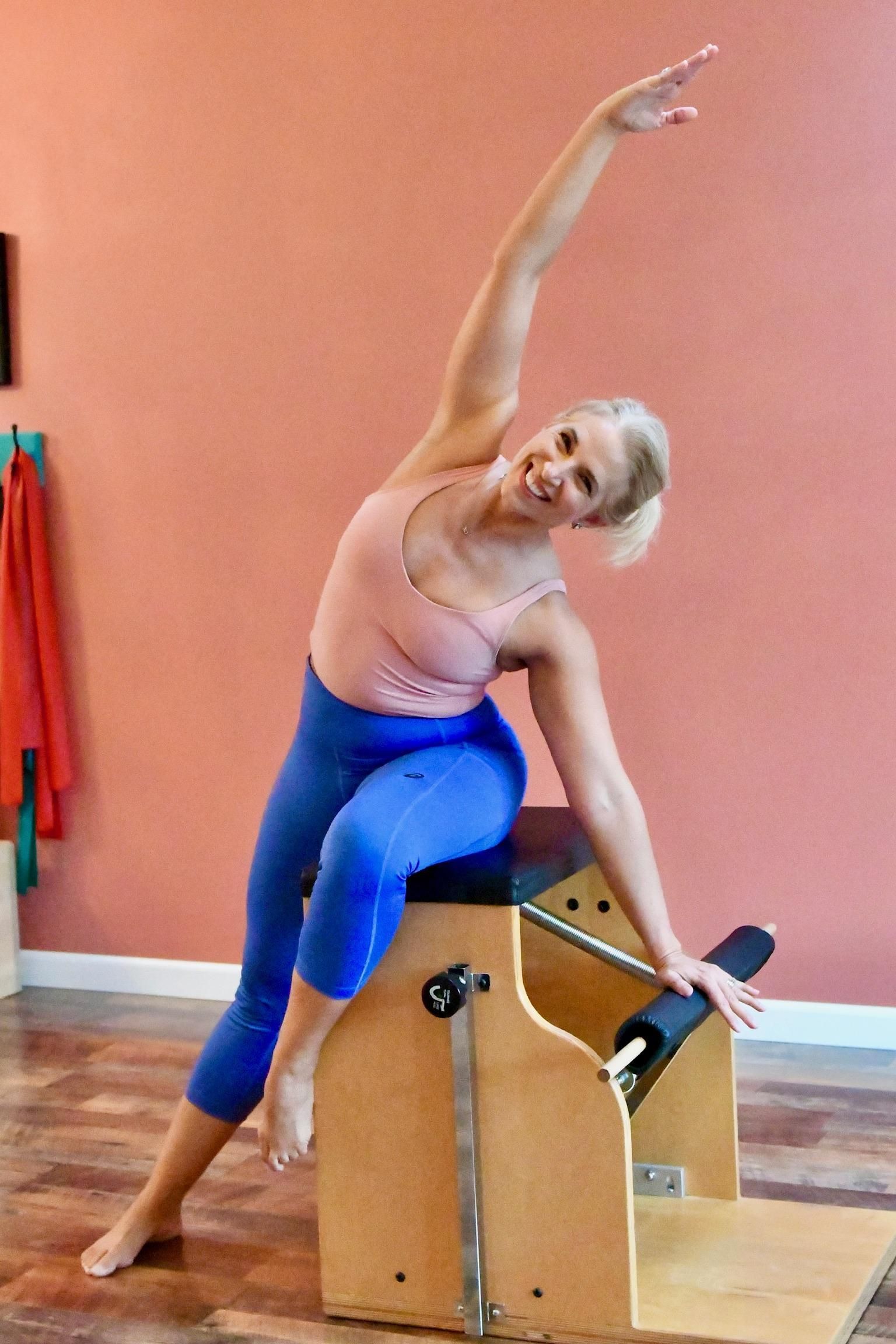 Woman in blue leggings and pink top doing Pilates on a wooden chair, arms extended.