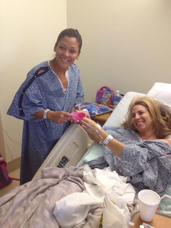 A woman in a hospital gown is giving a flower to another woman in a hospital bed.