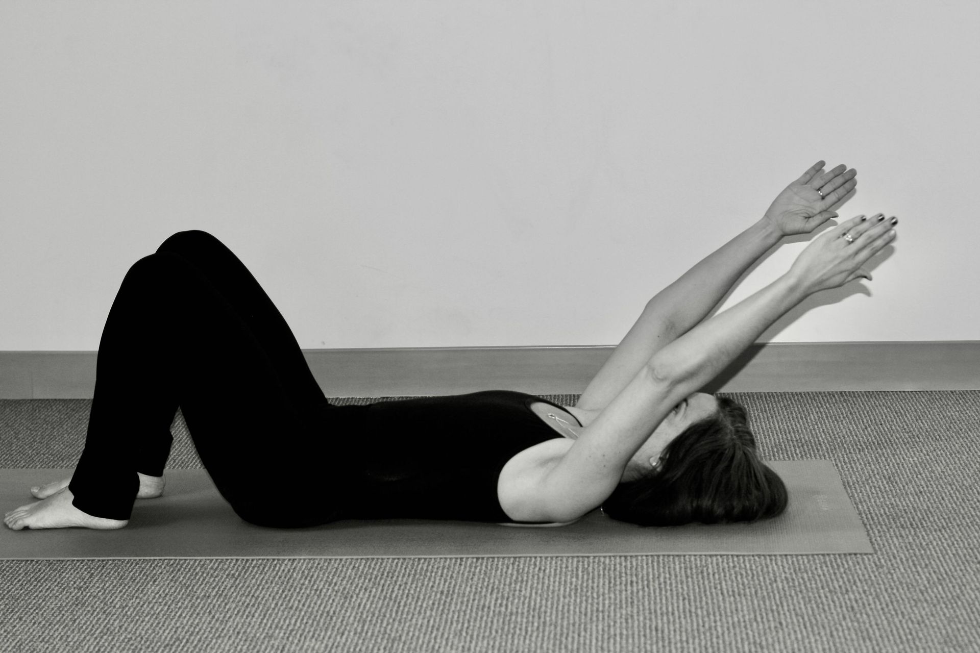 Person lying on a yoga mat, knees bent, reaching arms overhead during a floor exercise.