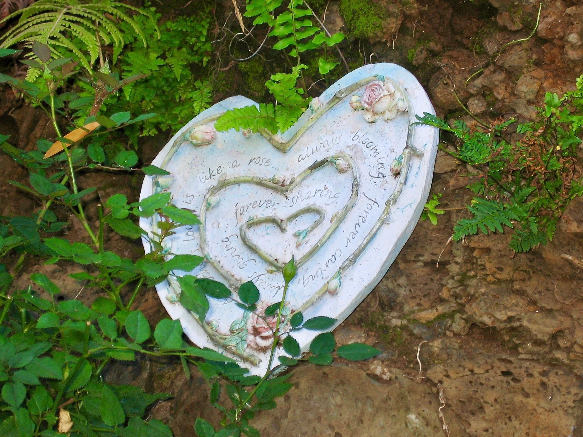 A white heart shaped plaque is sitting on the ground surrounded by plants.