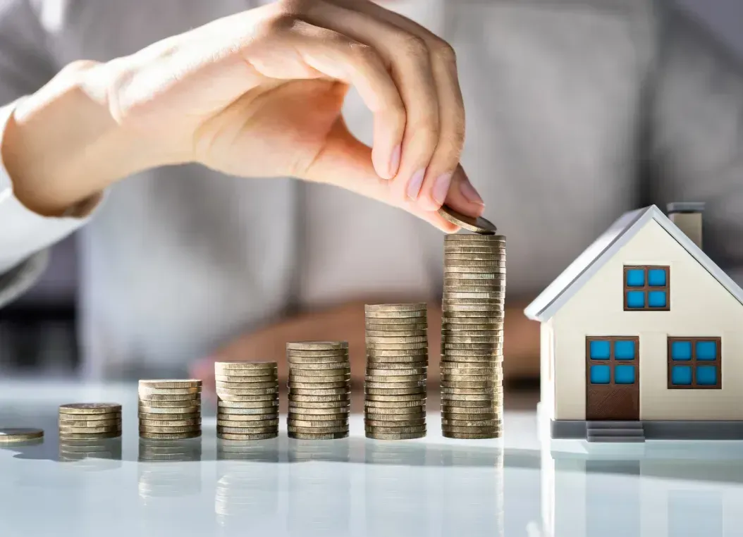 A person is putting coins into a stack next to a model house.