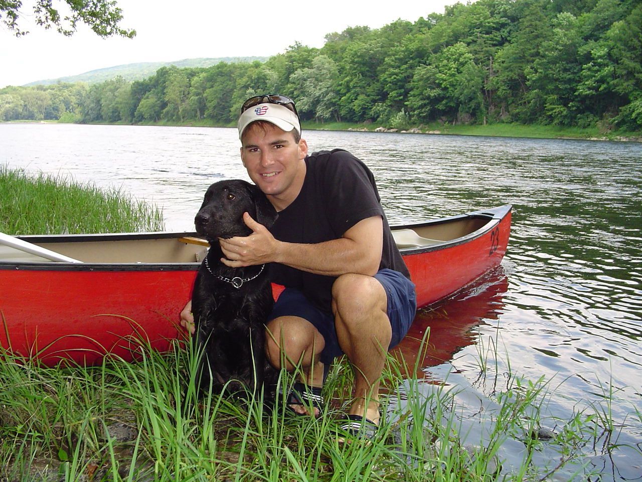 A man kneeling next to a red adventure sports canoe holding a black dog