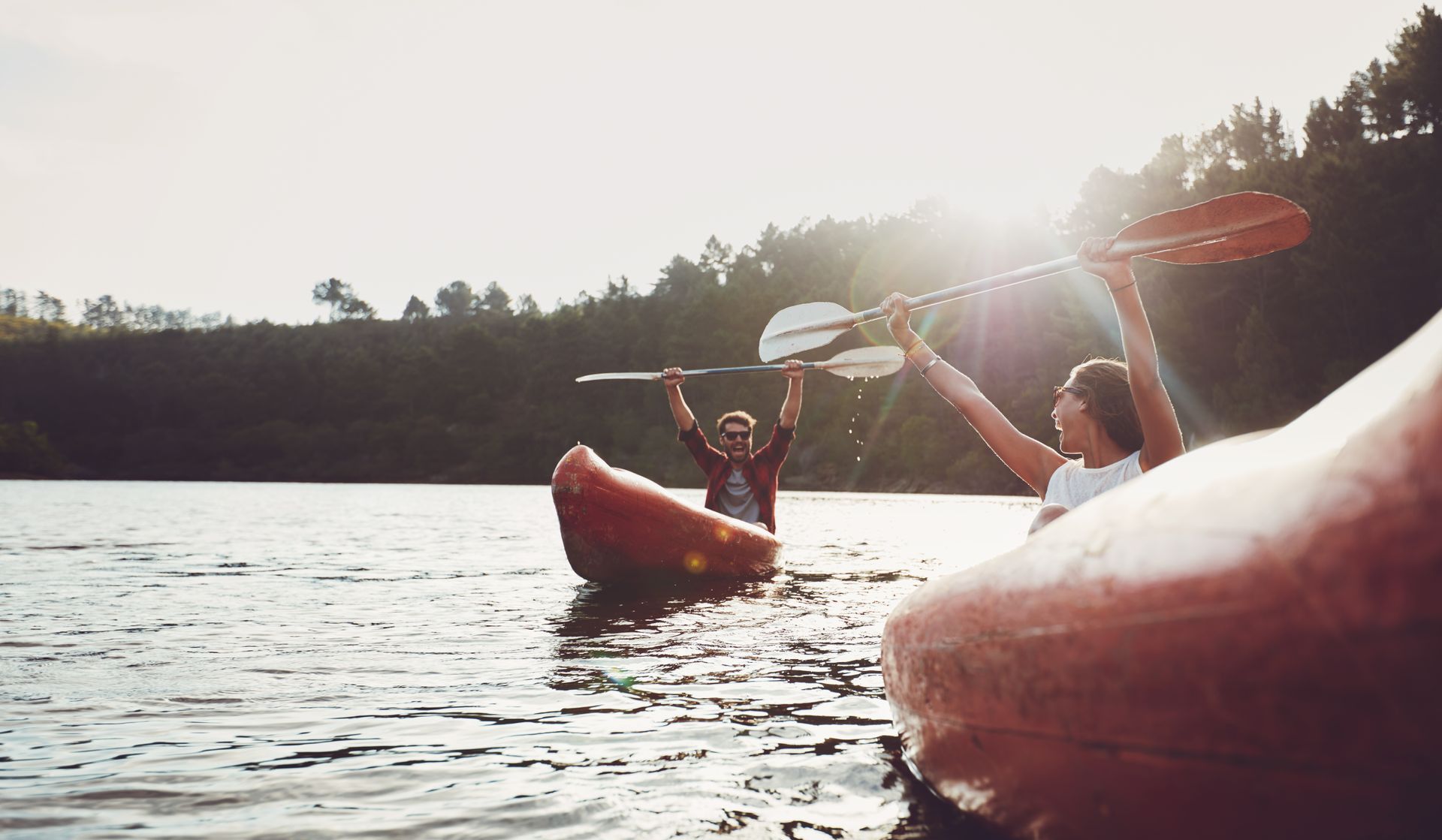 Two people kayaking on a lake, raising paddles in the sunlight near a forested shore.