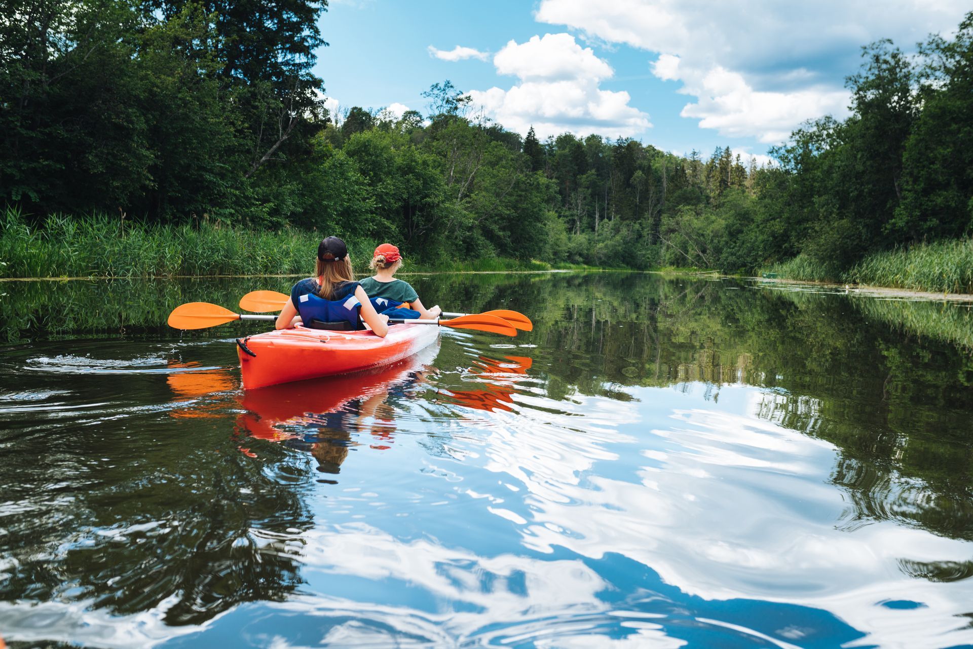 Two people kayaking in a red canoe on a calm river, surrounded by trees and blue sky.
