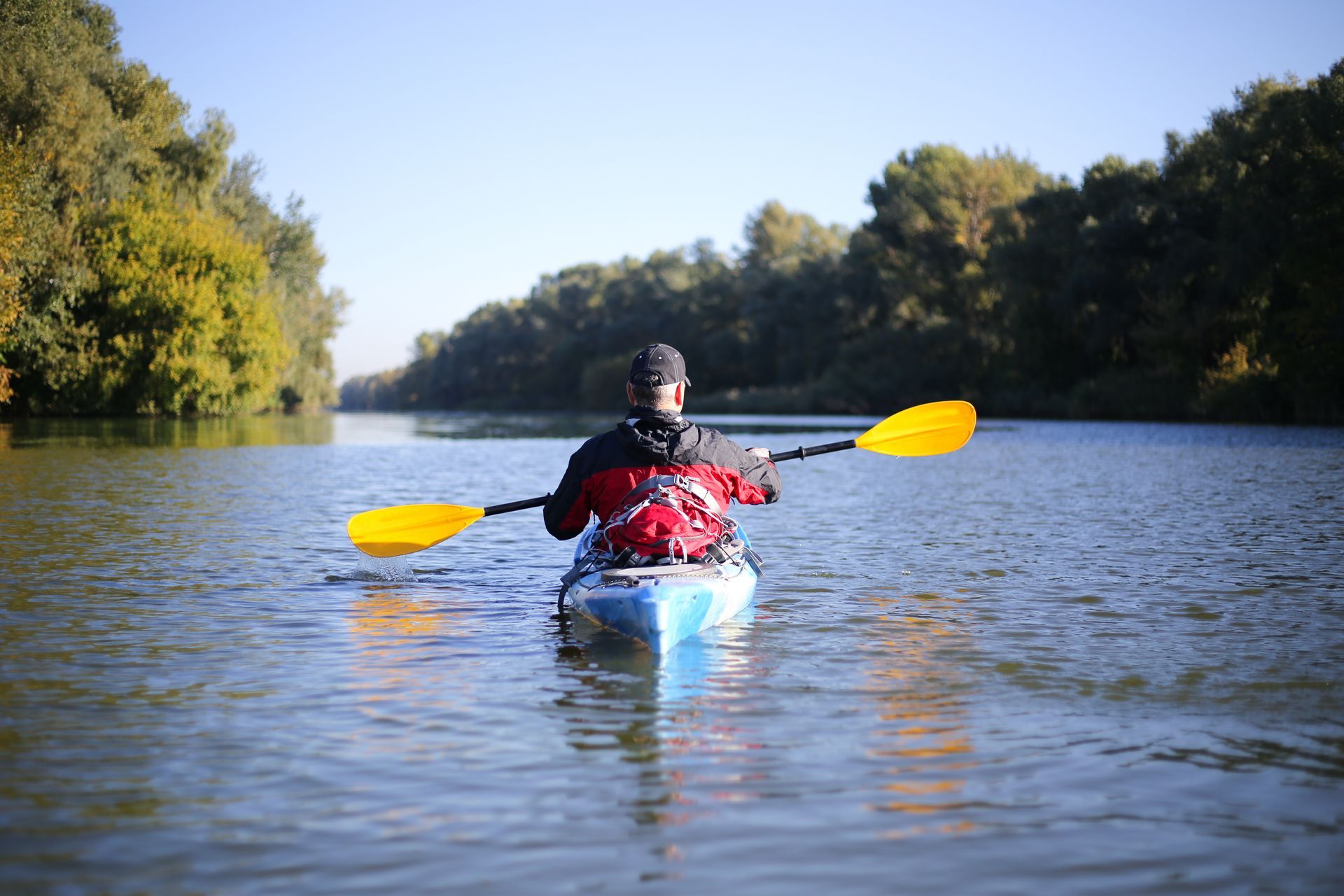 Kayaker in a blue canoe paddles on a calm river lined with trees under a clear sky