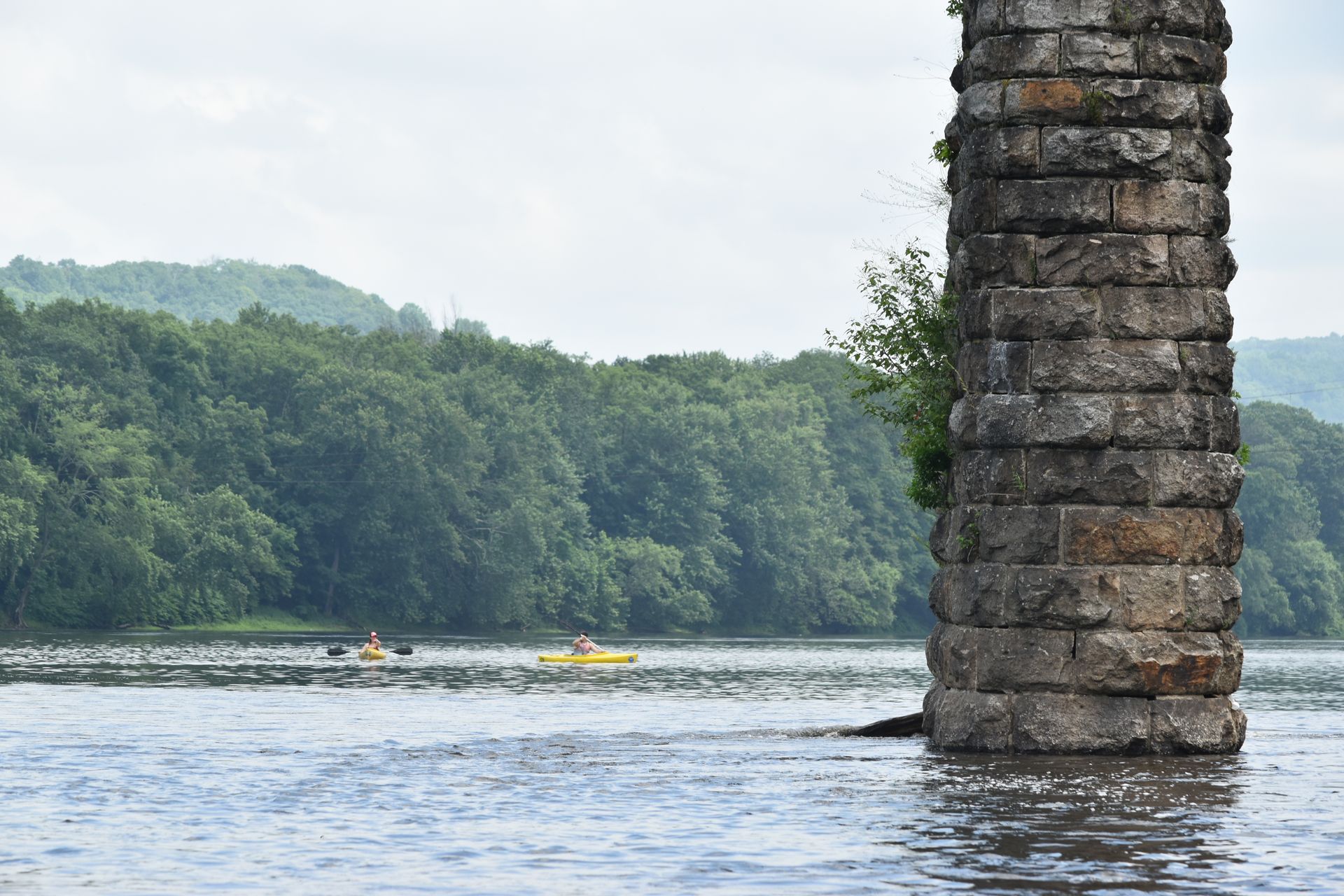 A person in a yellow adventure sports kayak is paddling under a bridge over a river.